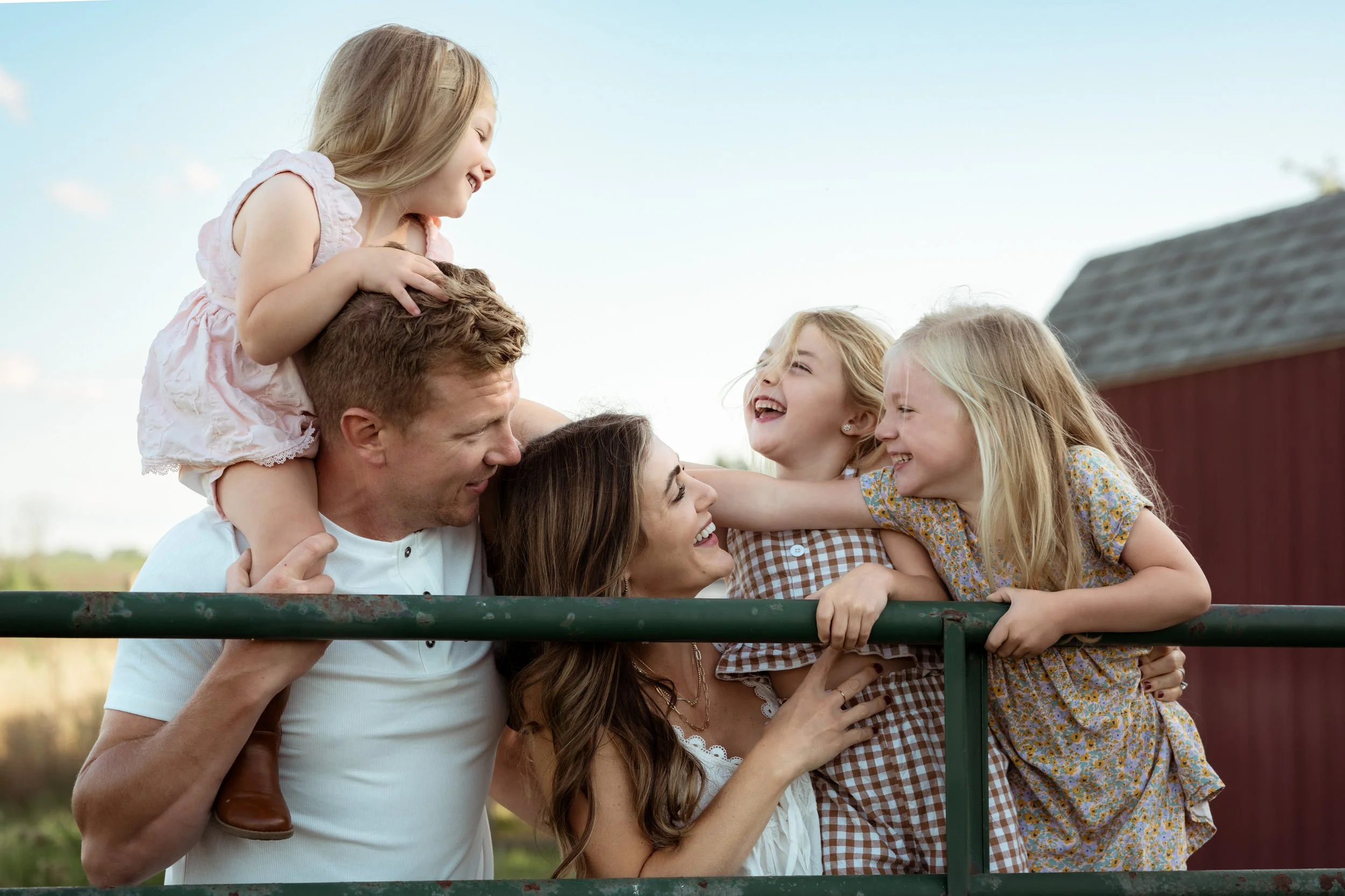 A family laughing together during an outdoor photoshoot, with a father holding his daughter on his shoulders and a mother embracing their two girls as they lean over a green farm gate. Soft golden light and a rustic barn setting create a joyful, cand