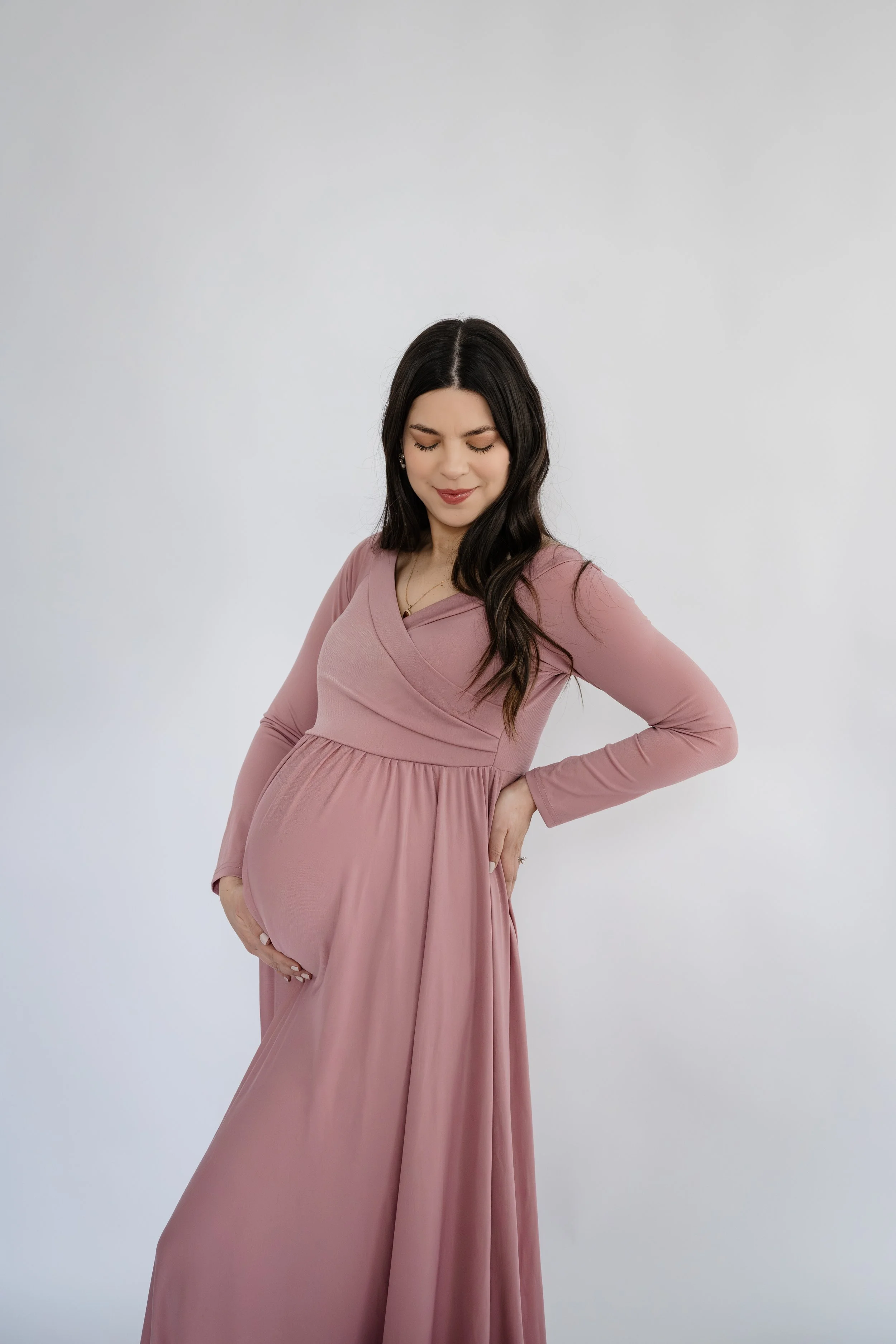 Pregnant woman in a flowing mauve maternity dress gently cradles her baby bump during a minimalist studio maternity session against a soft white background.