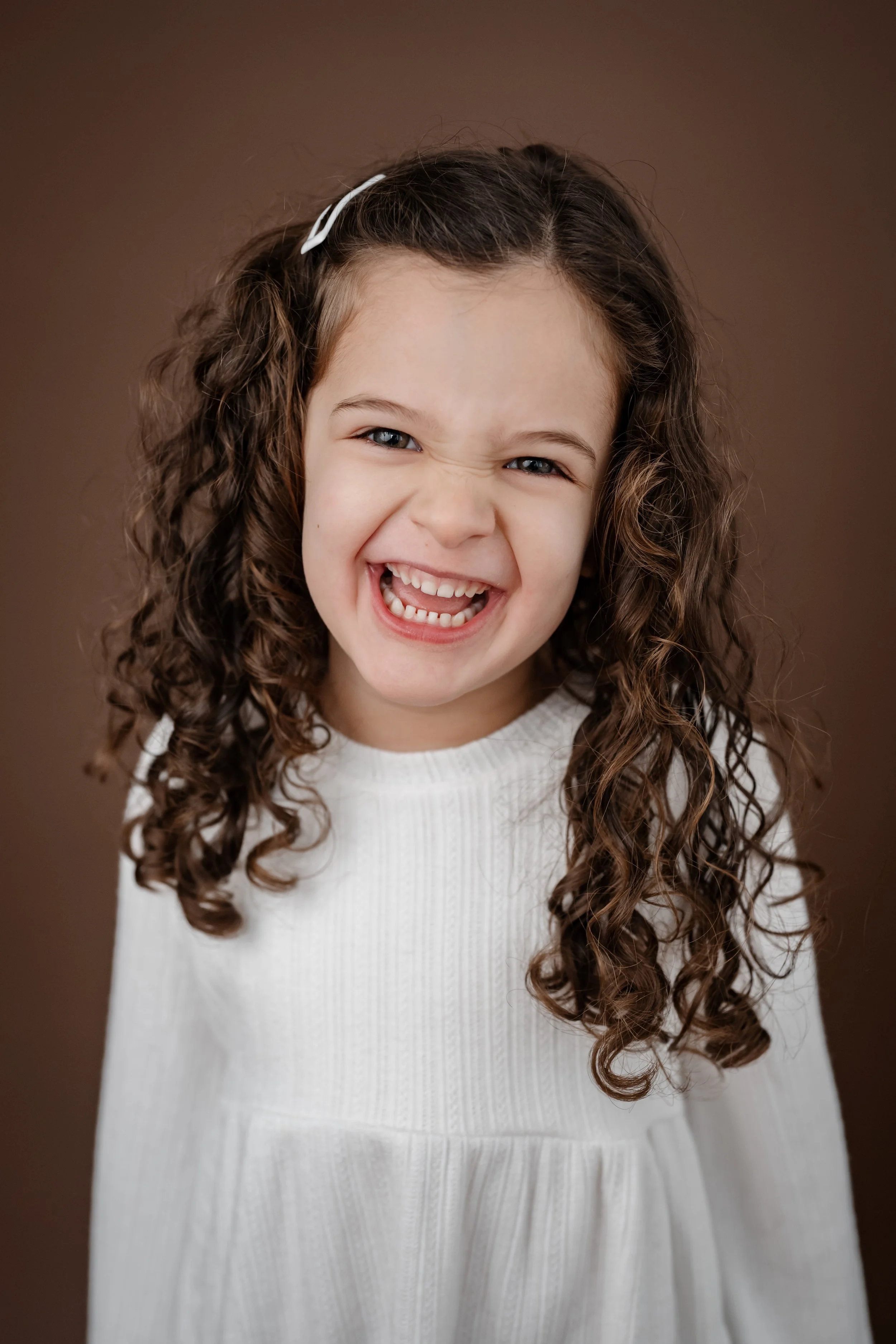 Young girl with curly hair laughs joyfully during a child portrait session, wearing a white dress against a warm brown studio backdrop.