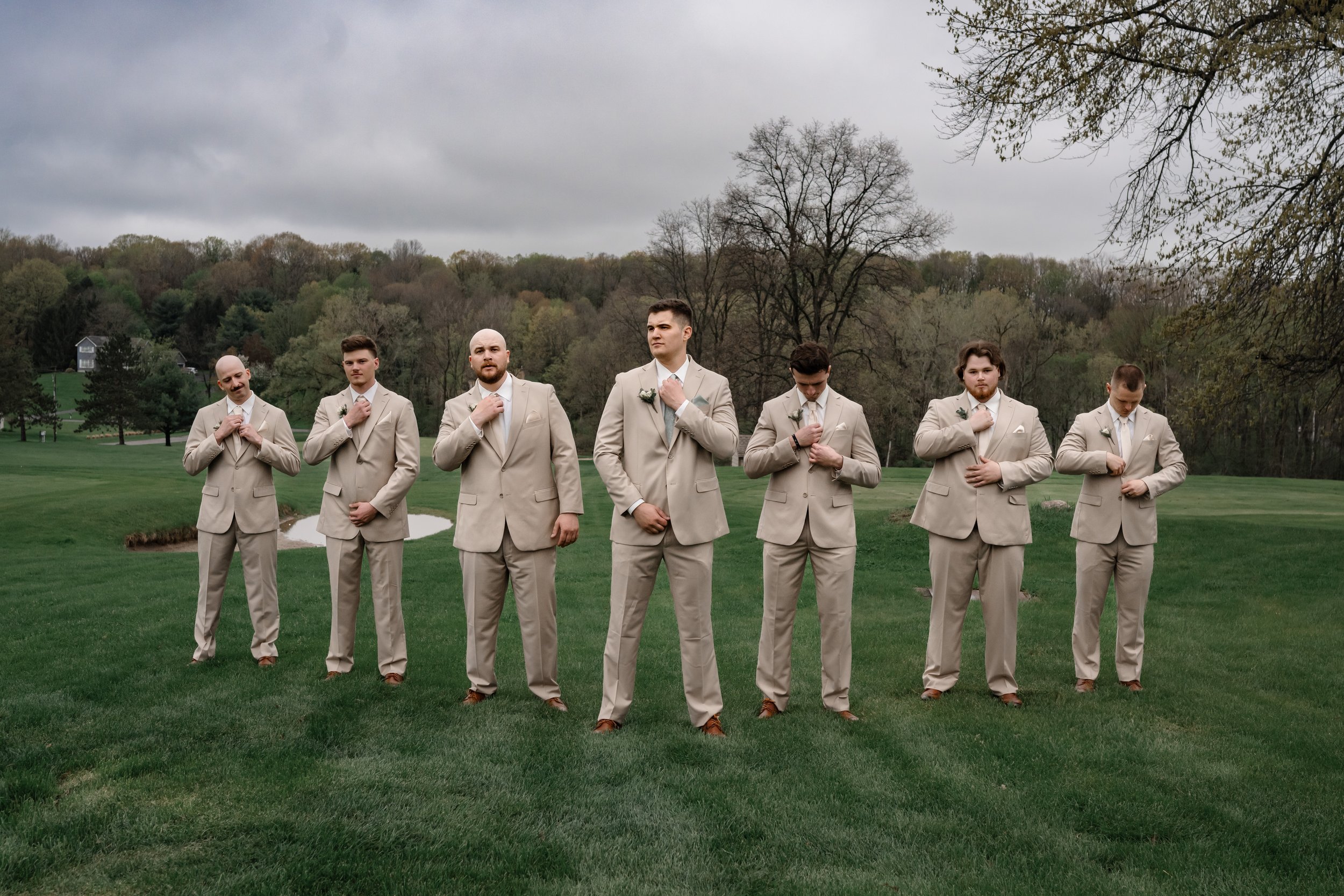 A group of groomsmen standing in a line outdoors, adjusting their tan suits during a wedding day portrait on a grassy landscape.