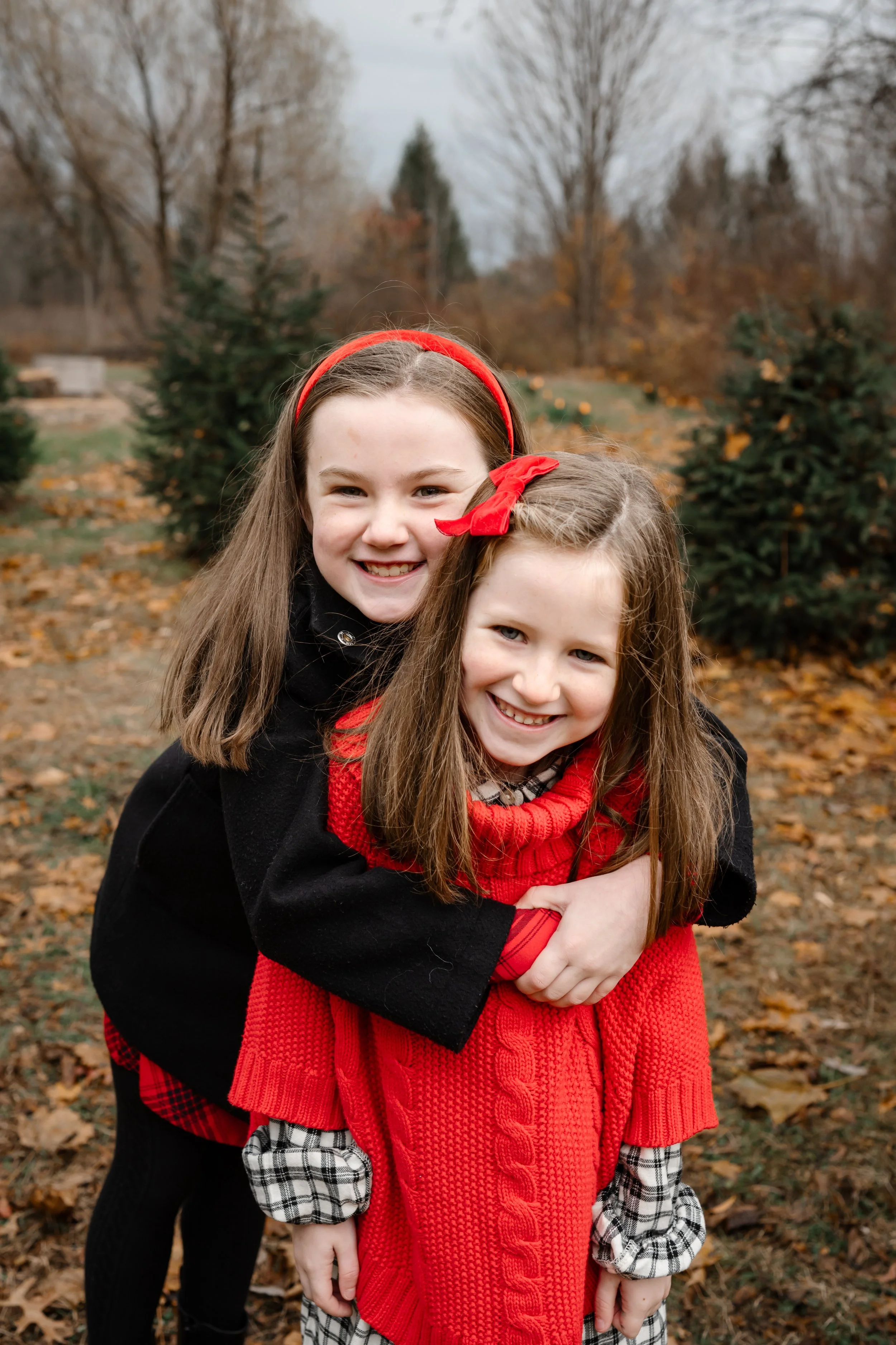 Two young sisters smile and hug outdoors during a fall family session, surrounded by autumn leaves and evergreen trees.