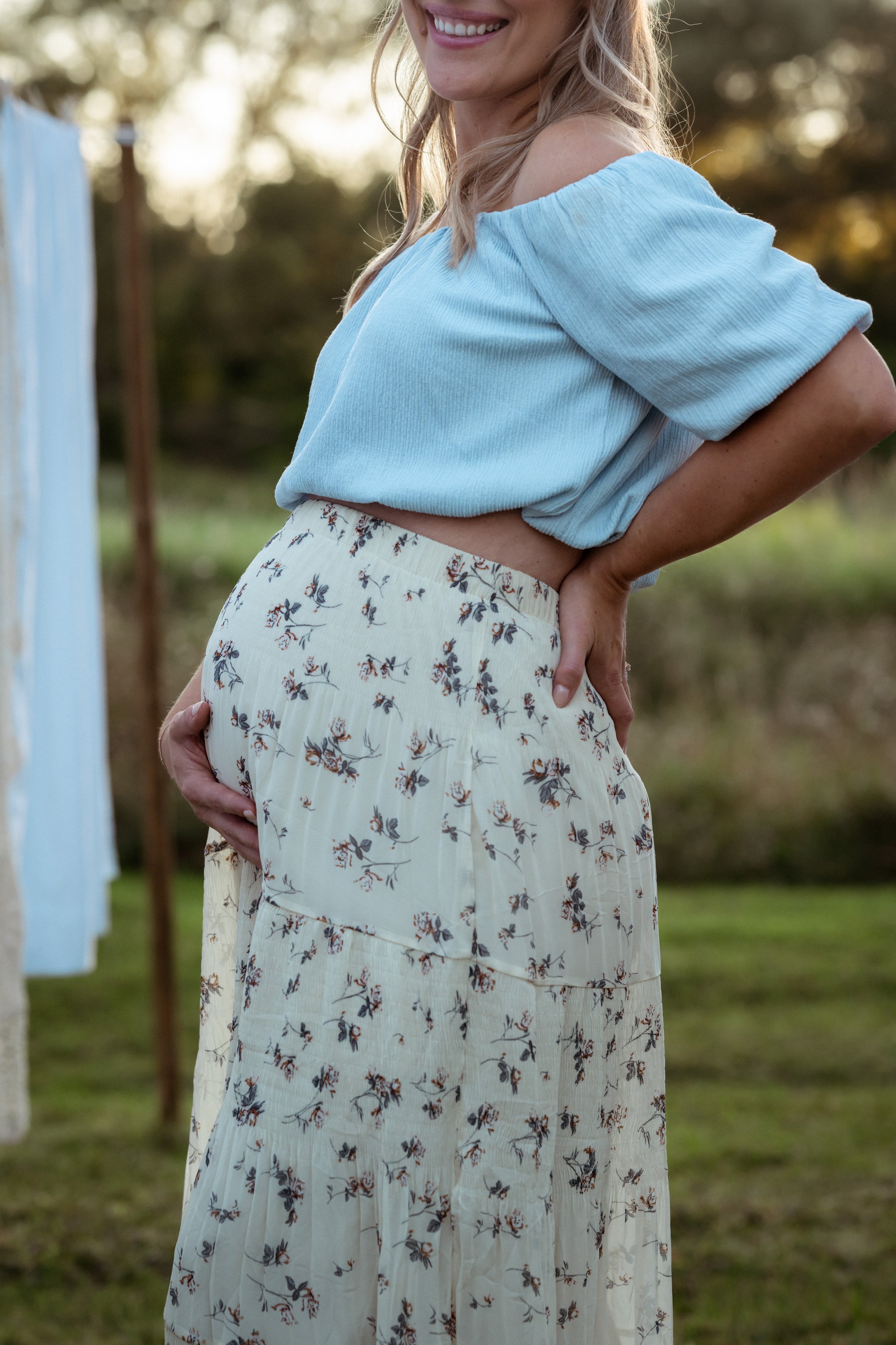 A pregnant woman standing outdoors at golden hour, gently holding her baby bump. She’s wearing a light blue off-the-shoulder top and a flowy floral skirt, smiling softly as the sunlight glows in the background.