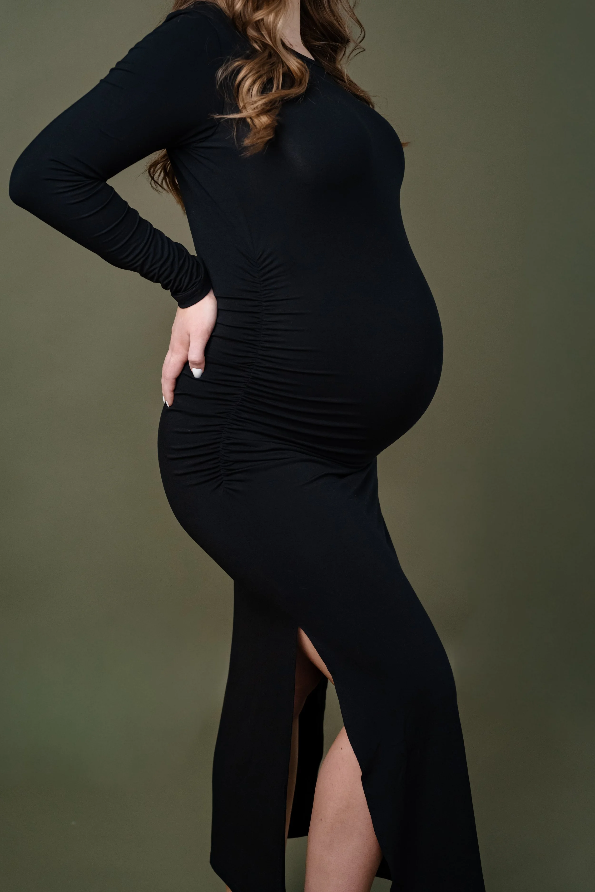 Side profile of a pregnant woman in a fitted black dress, gently holding her lower back and highlighting her baby bump against a soft neutral backdrop.