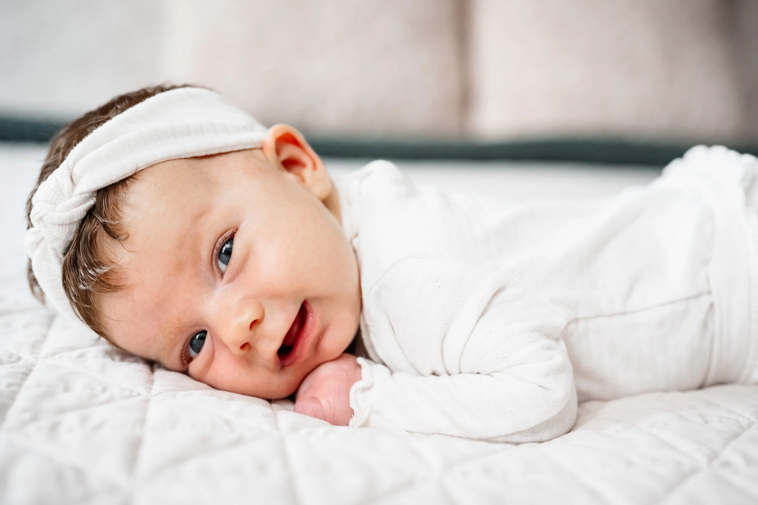Close-up of a newborn baby girl lying on a bed in soft natural light, wearing a white outfit and headband.