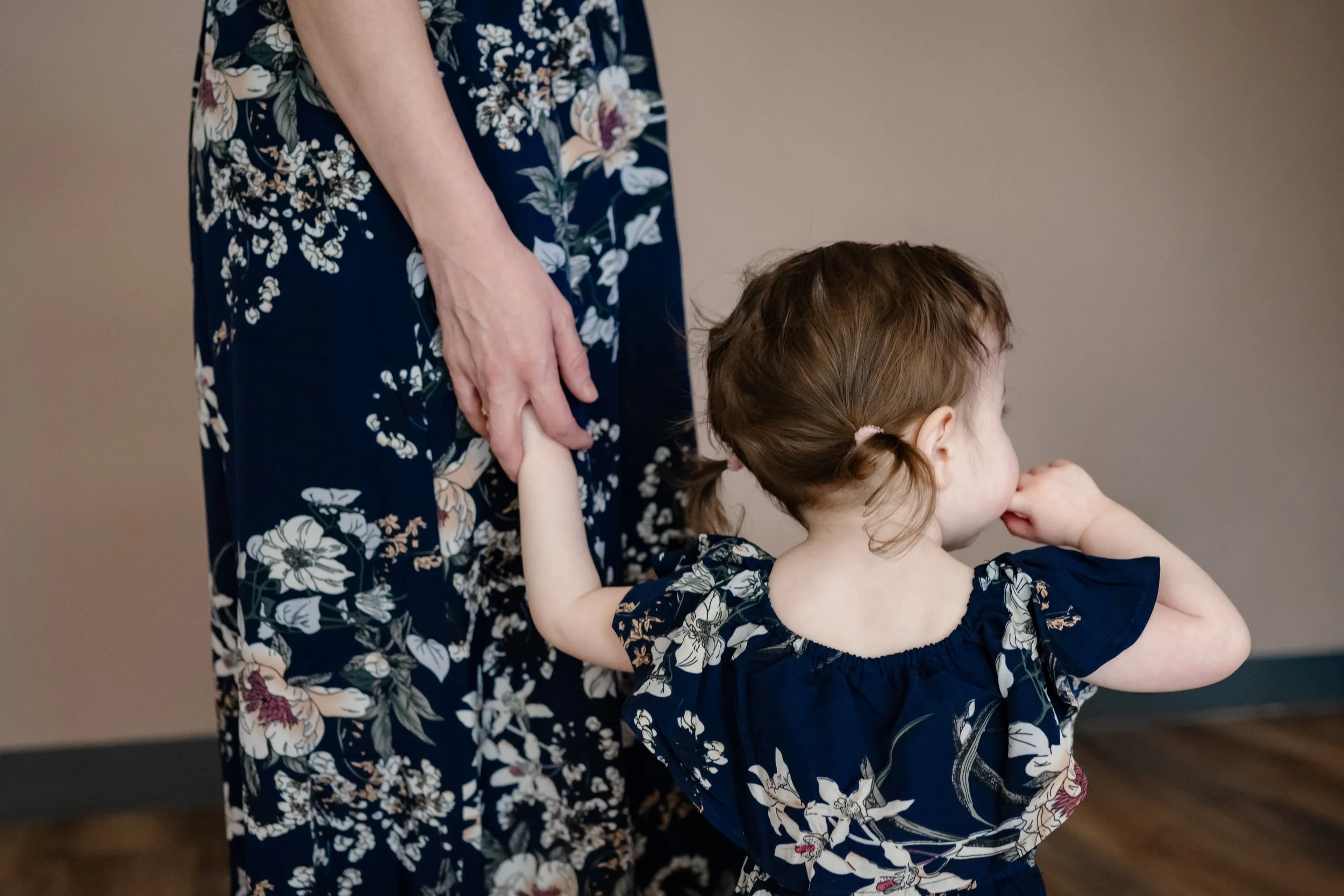 Close-up of a young child in a navy floral dress holding an adult’s hand, gently standing at their side, capturing a quiet, connected moment between parent and child.