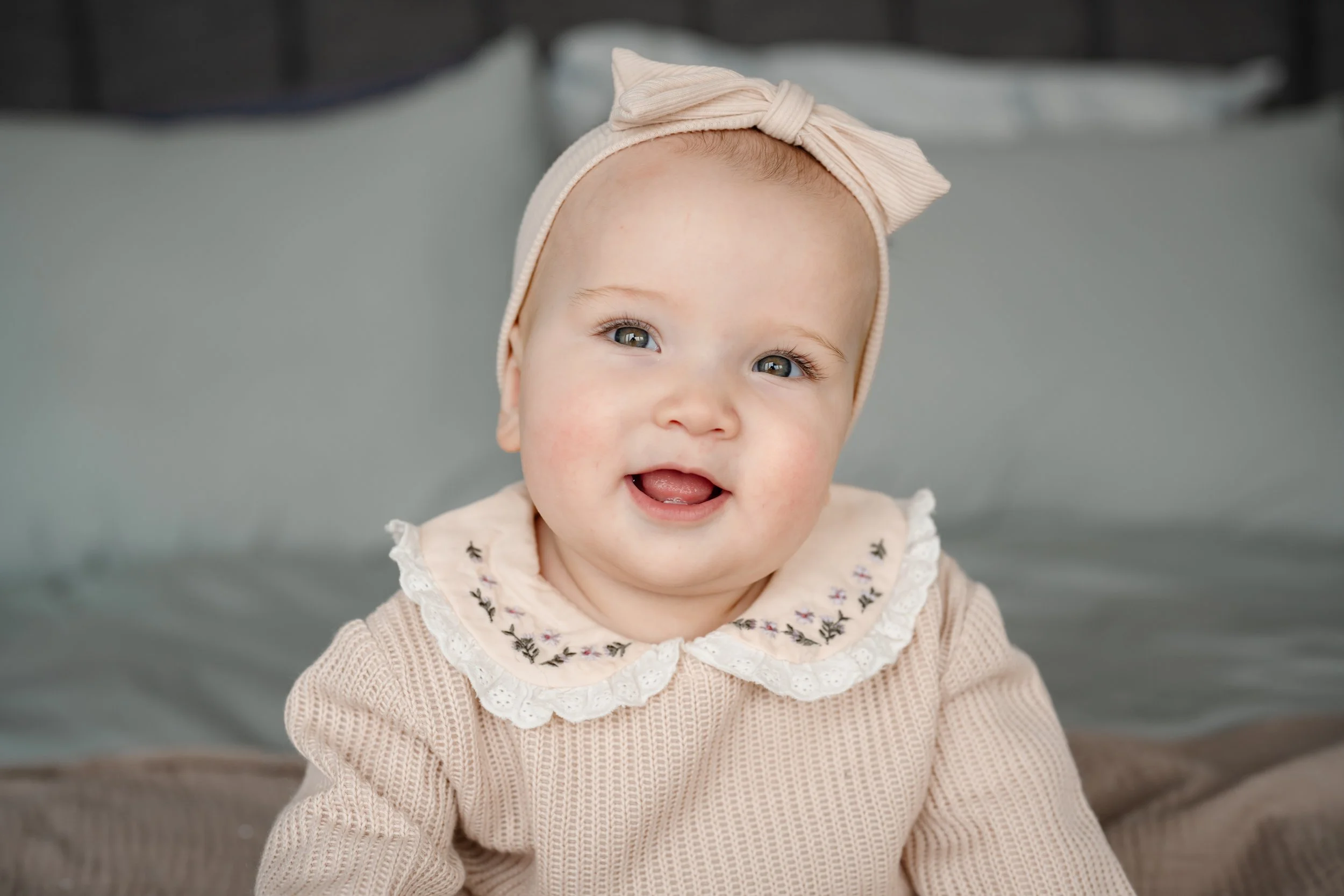 Smiling baby wearing a soft beige outfit with a lace collar and matching bow headband, sitting on a bed in a cozy, softly lit room.