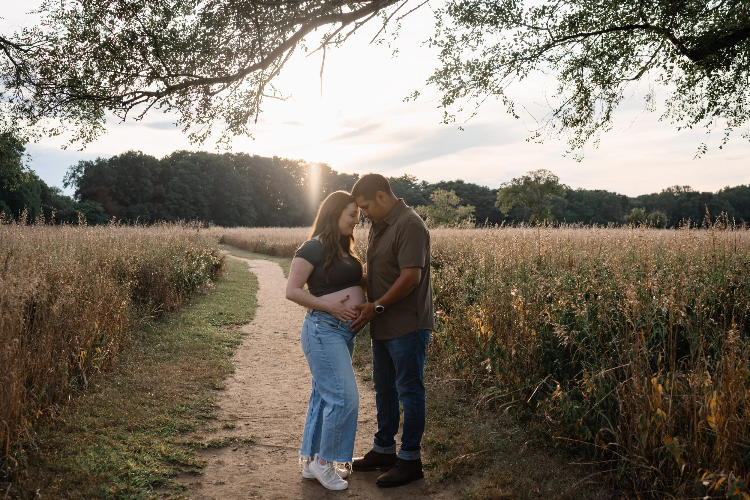 Expecting couple embracing on a sunlit path through a field during an outdoor maternity photography session in Rochester, NY.