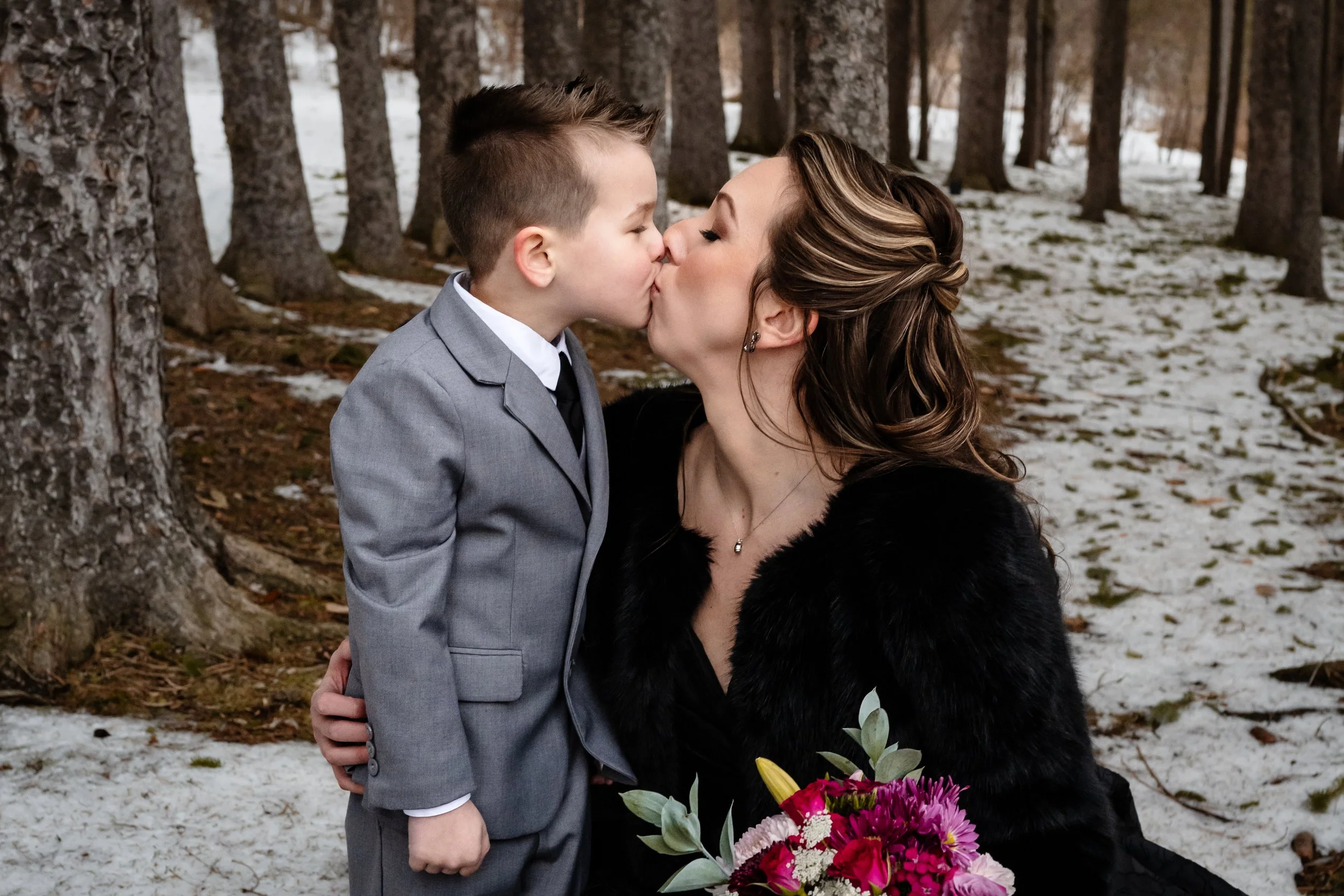 A bride sharing a sweet kiss with a young child outdoors in a winter setting, holding a colorful bouquet while surrounded by trees and patches of snow.