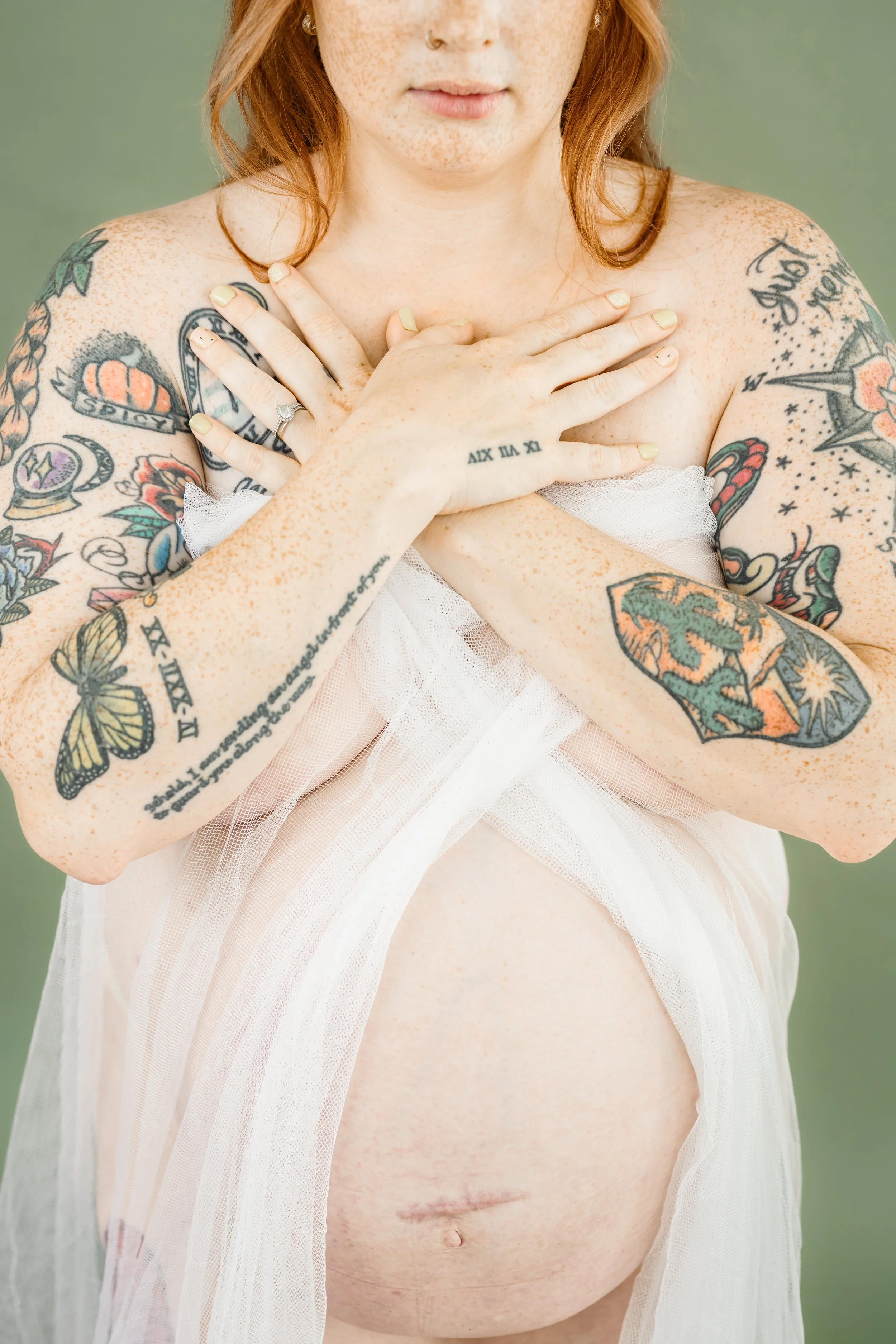 Close-up artistic maternity portrait of a tattooed pregnant woman draped in sheer fabric, showcasing her baby bump and C-section scar.