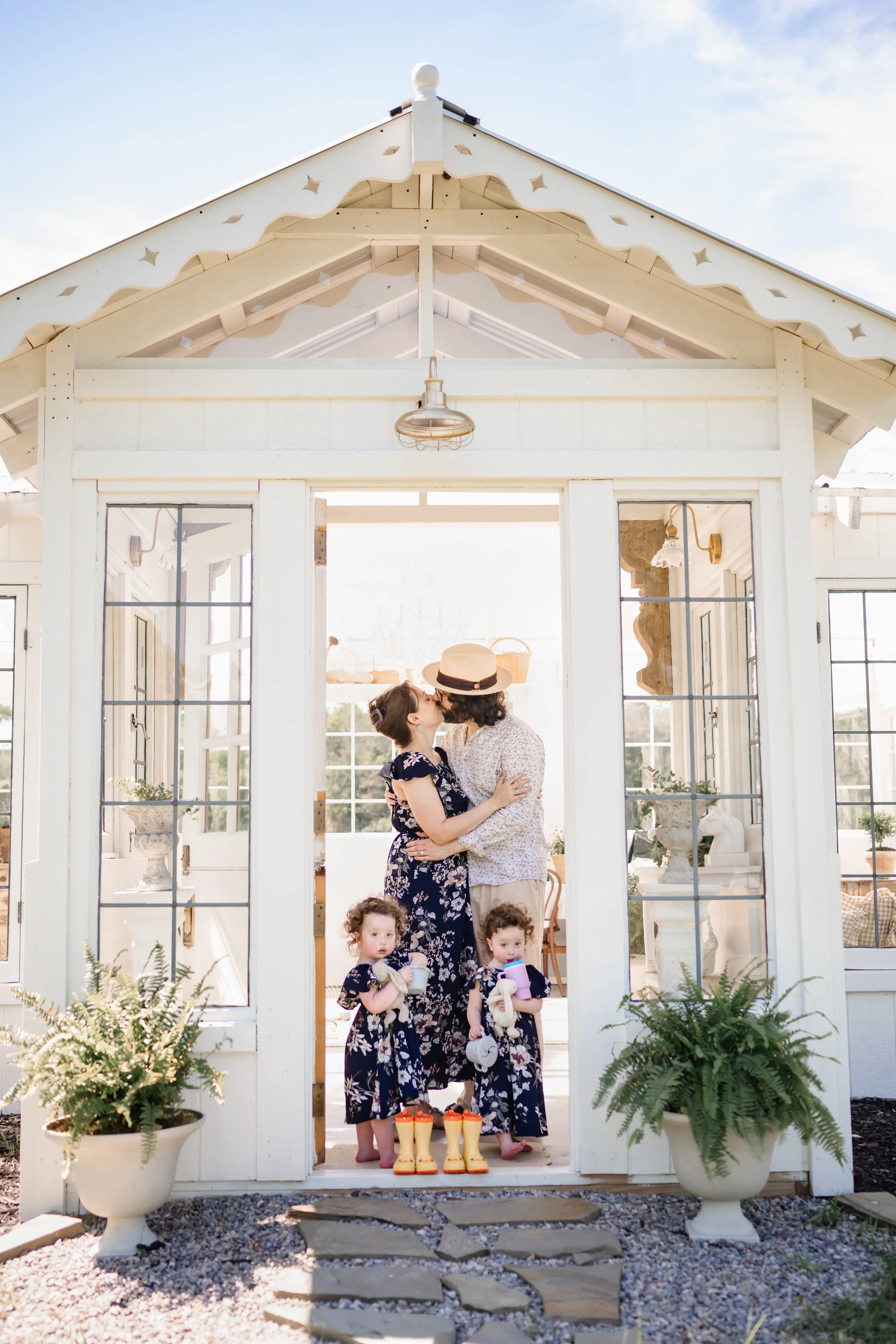 A family of four stands in the doorway of a bright white cottage as the parents share a kiss. Their two young daughters stand in front holding stuffed bunnies, with matching yellow rain boots lined up at their feet, creating a warm, storytelling fami