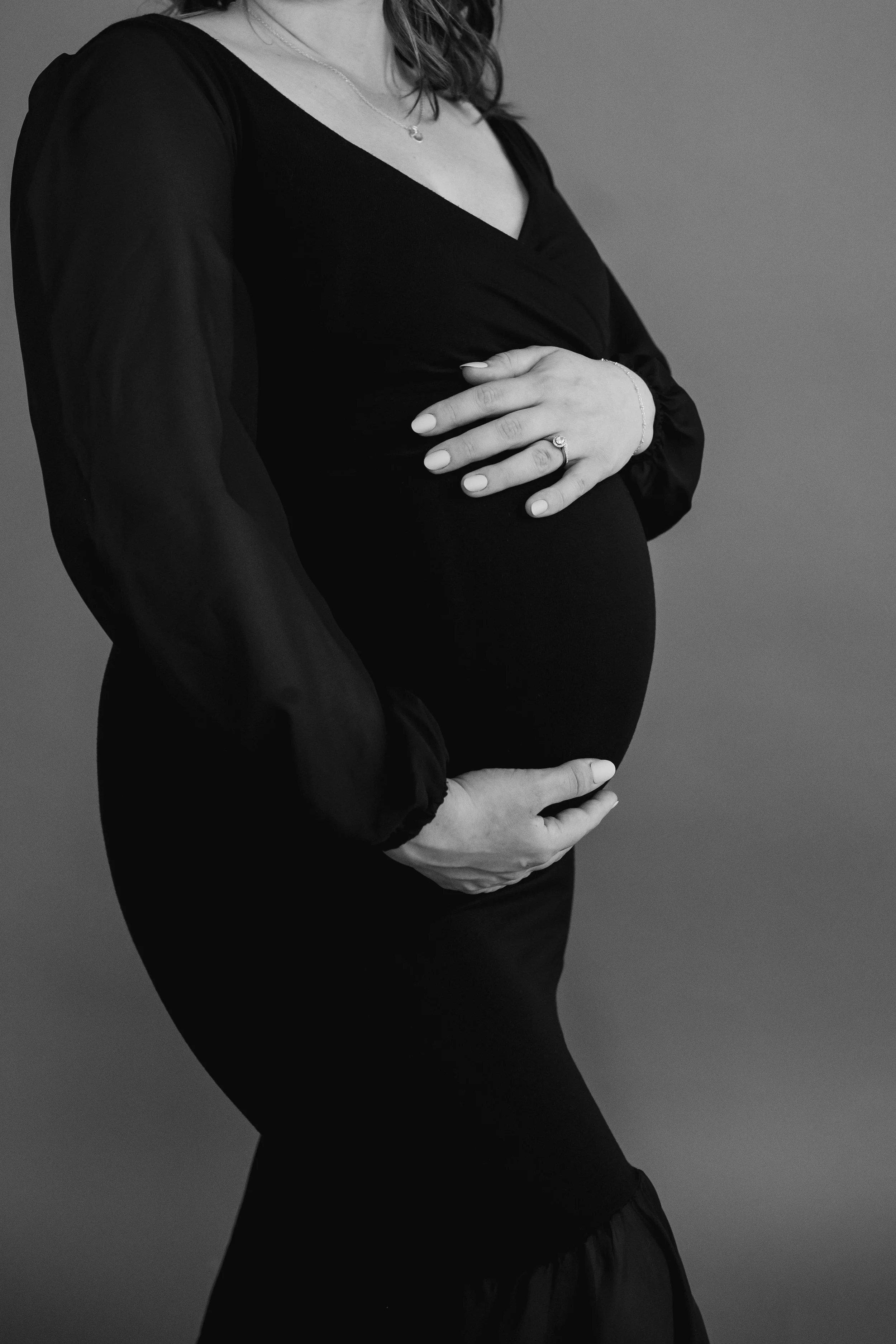 Black and white close-up of an expectant mother in a fitted dress, gently cradling her baby bump with both hands, highlighting the curve of her belly in a soft, minimal studio setting.