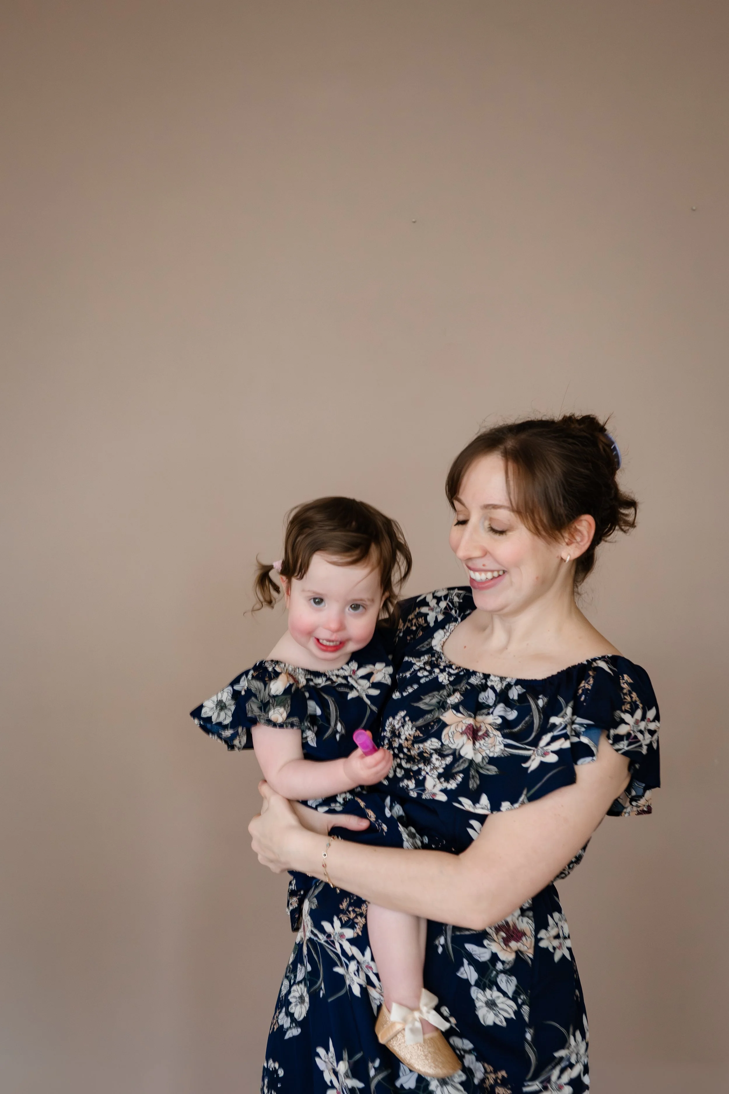 Mother smiles while holding her young daughter, who looks at the camera with a bright smile, during a studio family photography session against a neutral backdrop.