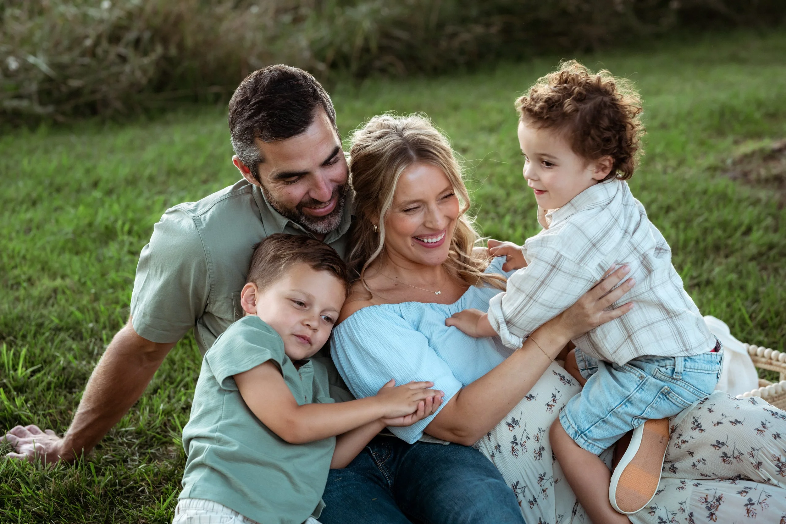 Happy family of four lying on green grass outdoors, smiling and playing together.