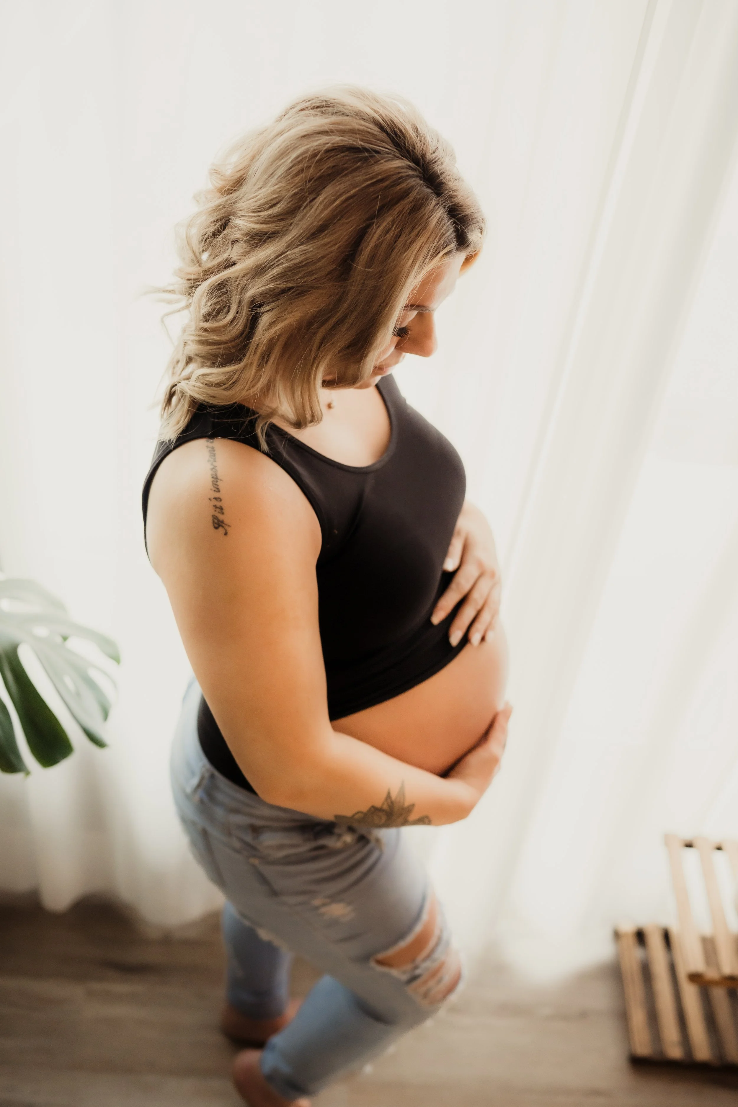 Light-filled maternity portrait of a pregnant woman standing by a window, gently holding her baby bump while wearing a black top and jeans in a soft, natural setting.