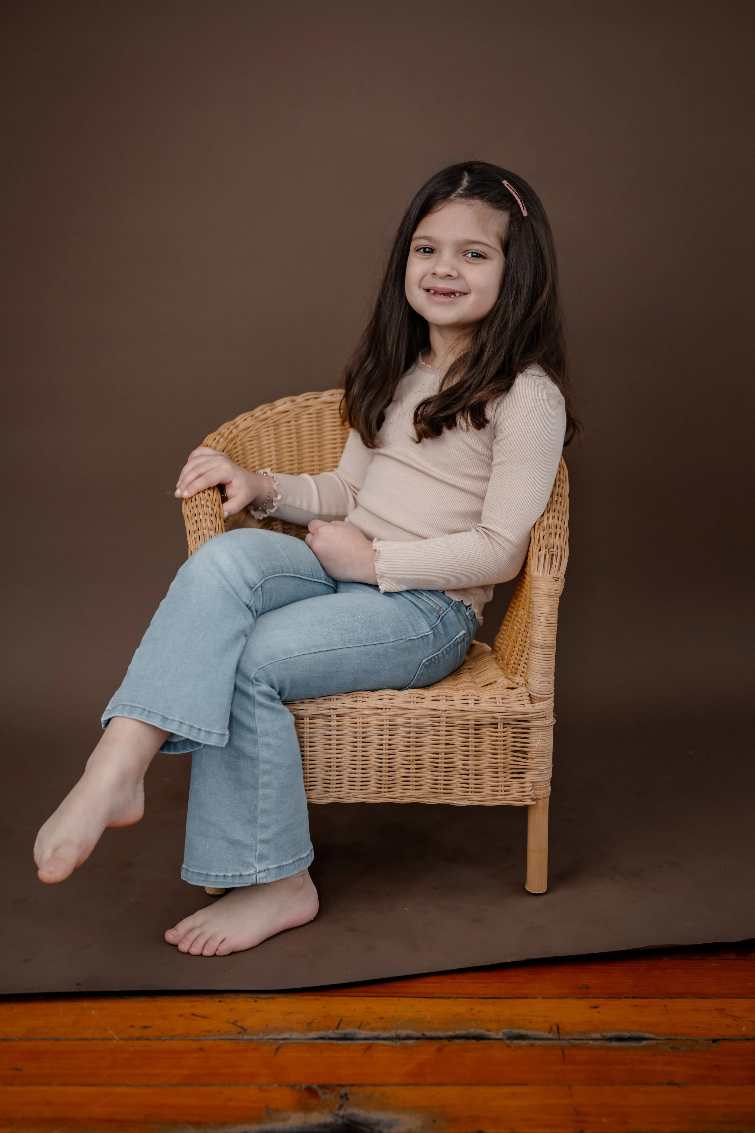 A young girl with long brown hair, wearing a beige long-sleeve top and light blue jeans, sitting barefoot on a wicker chair against a brown backdrop.