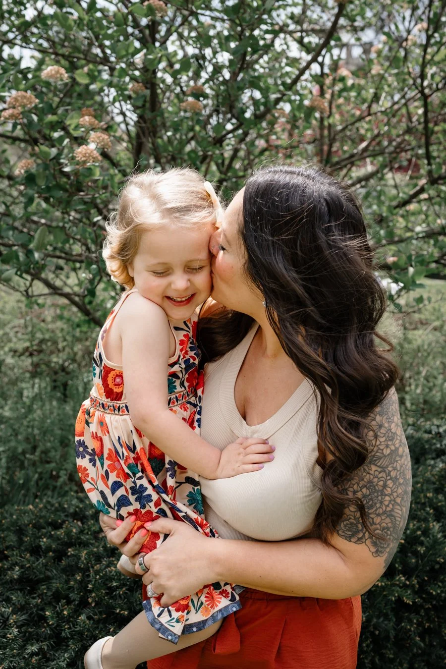 Mother holds her young daughter outdoors while kissing her cheek, capturing a joyful moment as the child smiles with eyes closed beneath flowering trees in soft natural light.