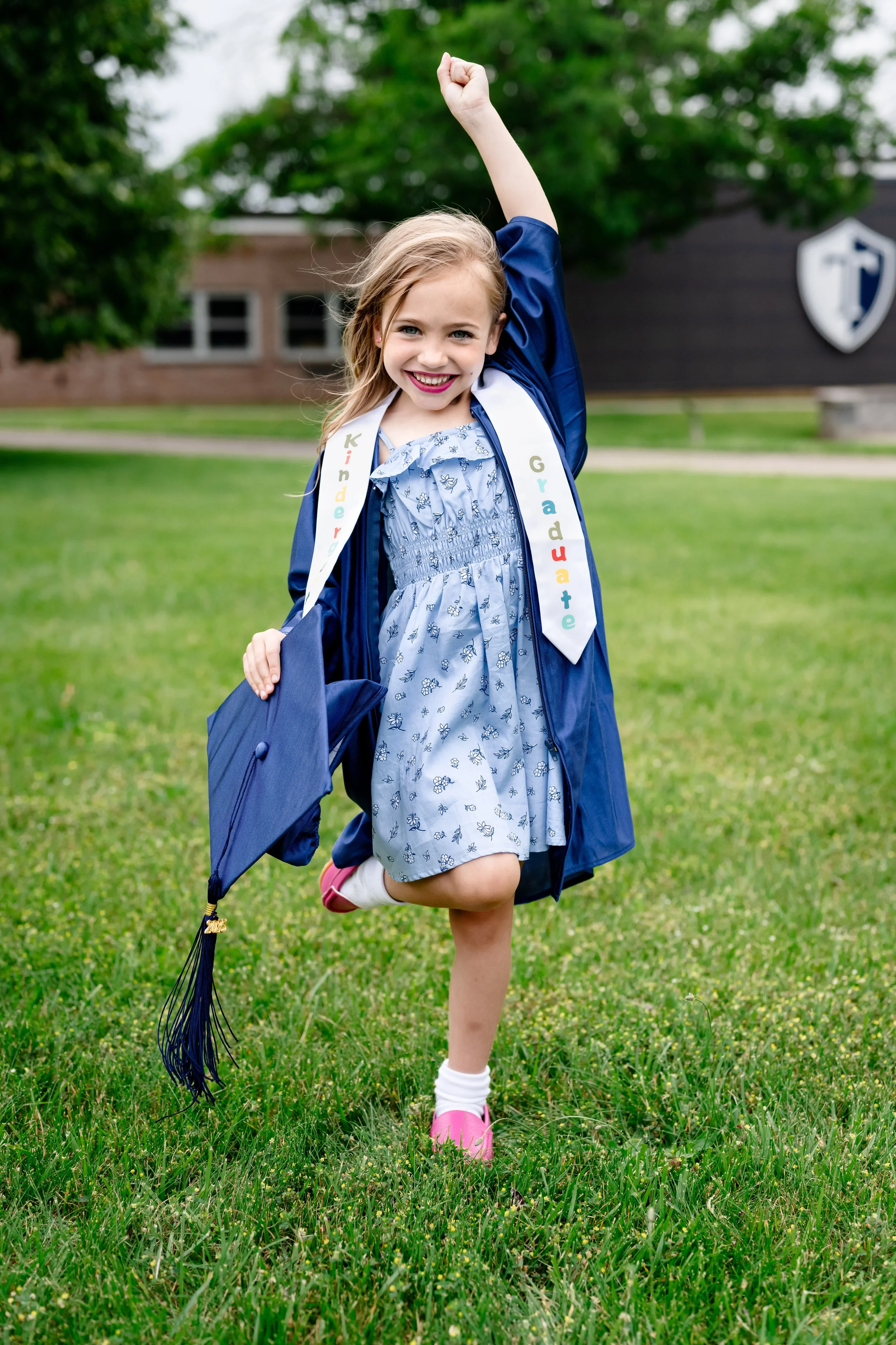 Child in a blue kindergarten graduation cap and gown stands on a grassy field, smiling with one arm raised in celebration while holding her cap, wearing a sash that reads “Kinder Graduate,” with a school building blurred in the background.