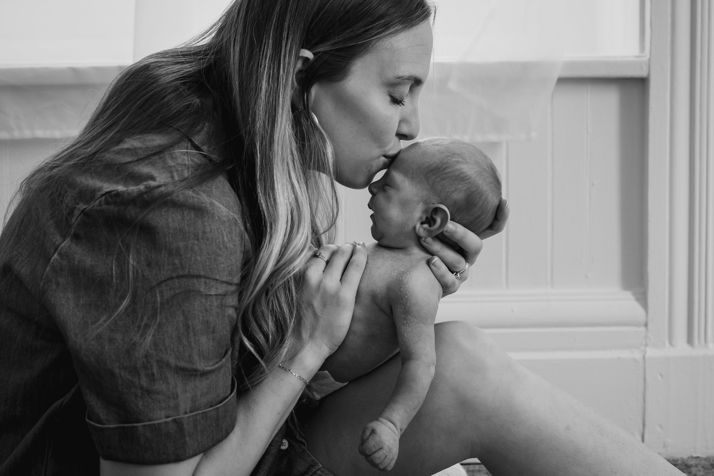 Black and white image of a mother gently kissing her newborn baby’s forehead while supporting their head, seated near a softly lit window.
