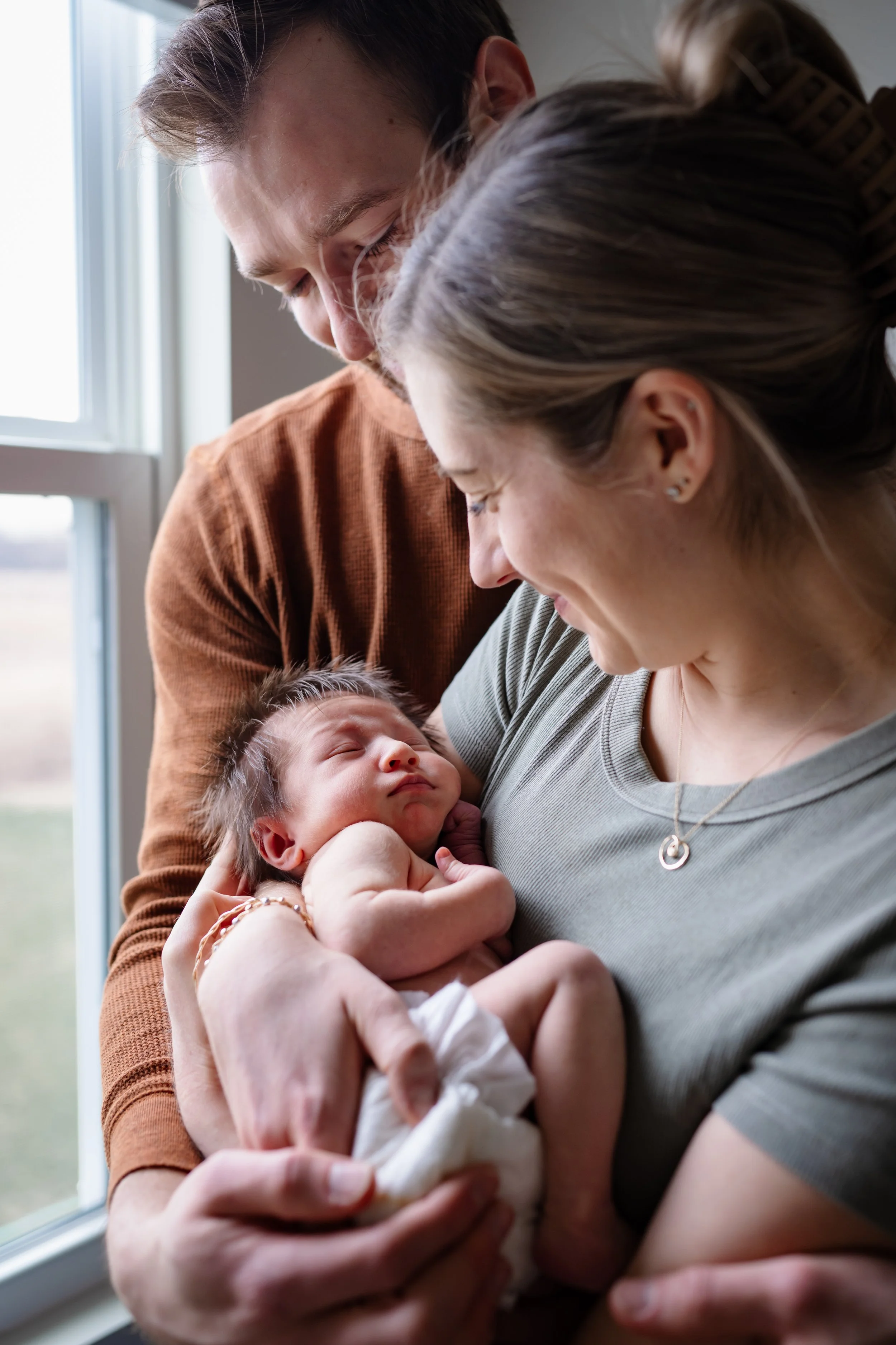 New parents smiling down at their newborn baby while holding them close near a softly lit window during an in-home session.