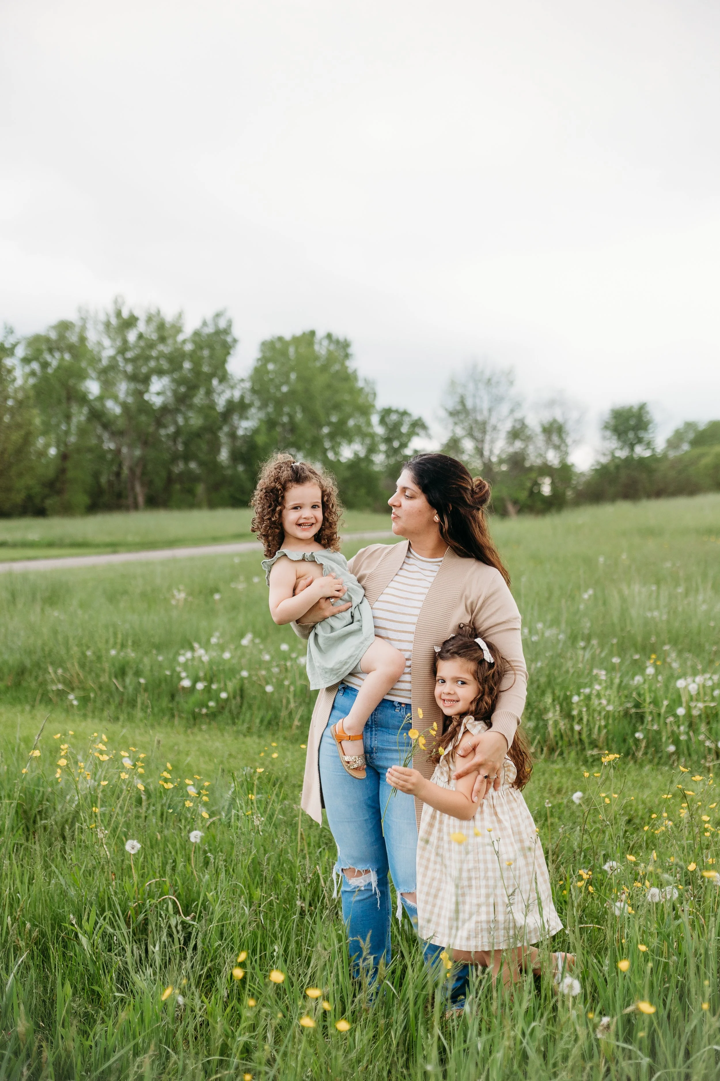 A woman holding a young girl in a green dress, standing in a field of wildflowers with another girl in a checkered dress holding yellow flowers, all smiling on a cloudy day.