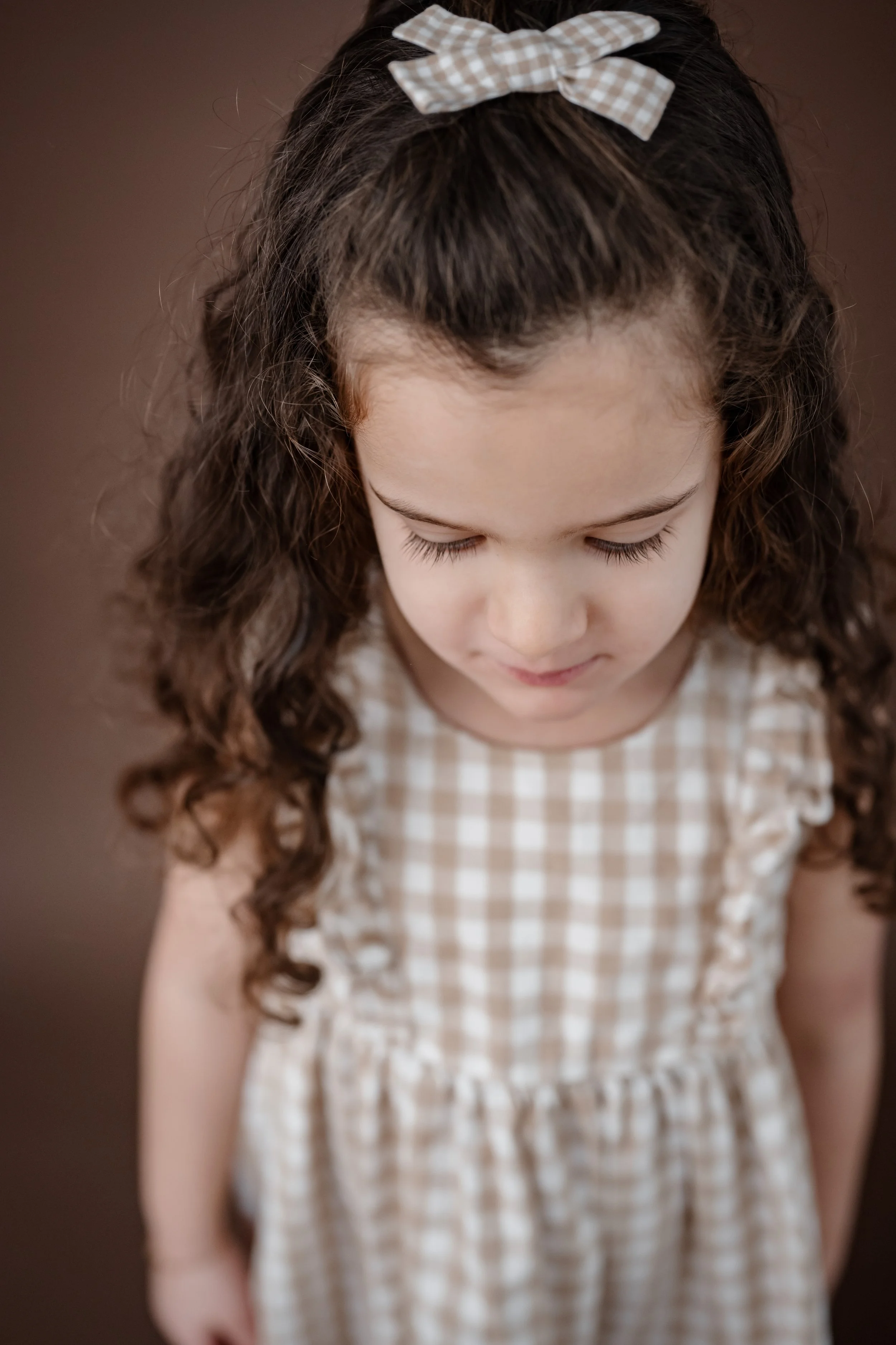 A young child looking down during a personality portrait session, wearing a gingham dress with curly hair and a bow, photographed against a warm brown backdrop.