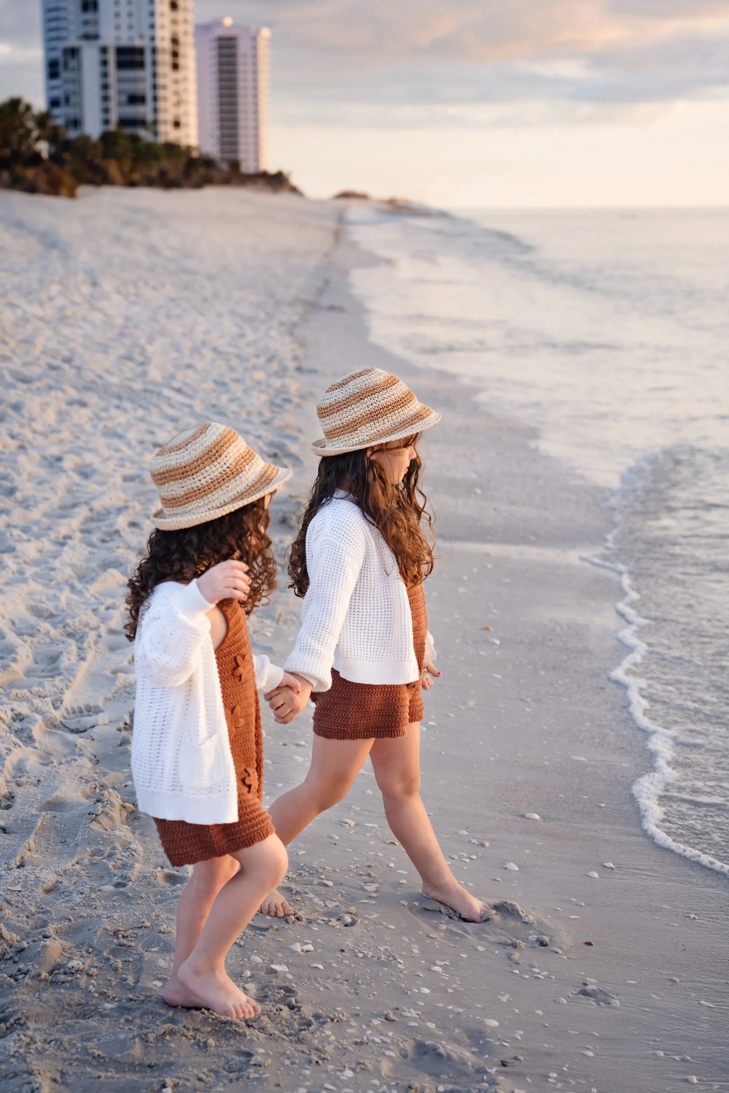 Two sisters walking along the beach in matching rust-colored outfits, white sweaters, and coordinating straw hats.