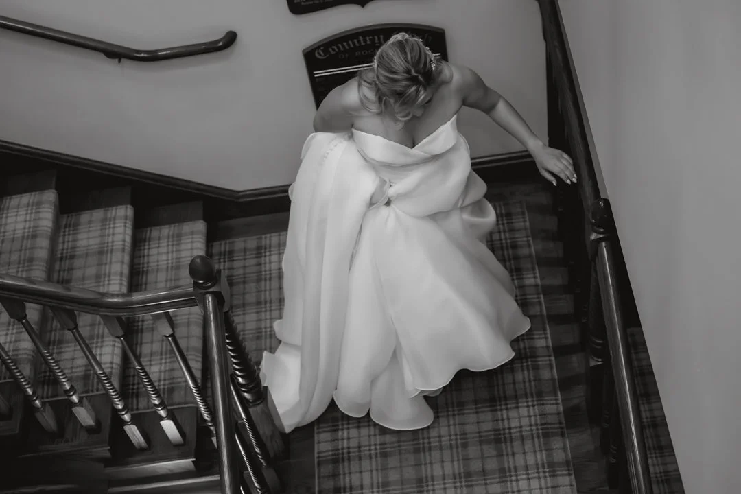 A black-and-white overhead view of a bride walking down a staircase, holding her wedding gown as it flows behind her in a classic interior setting.