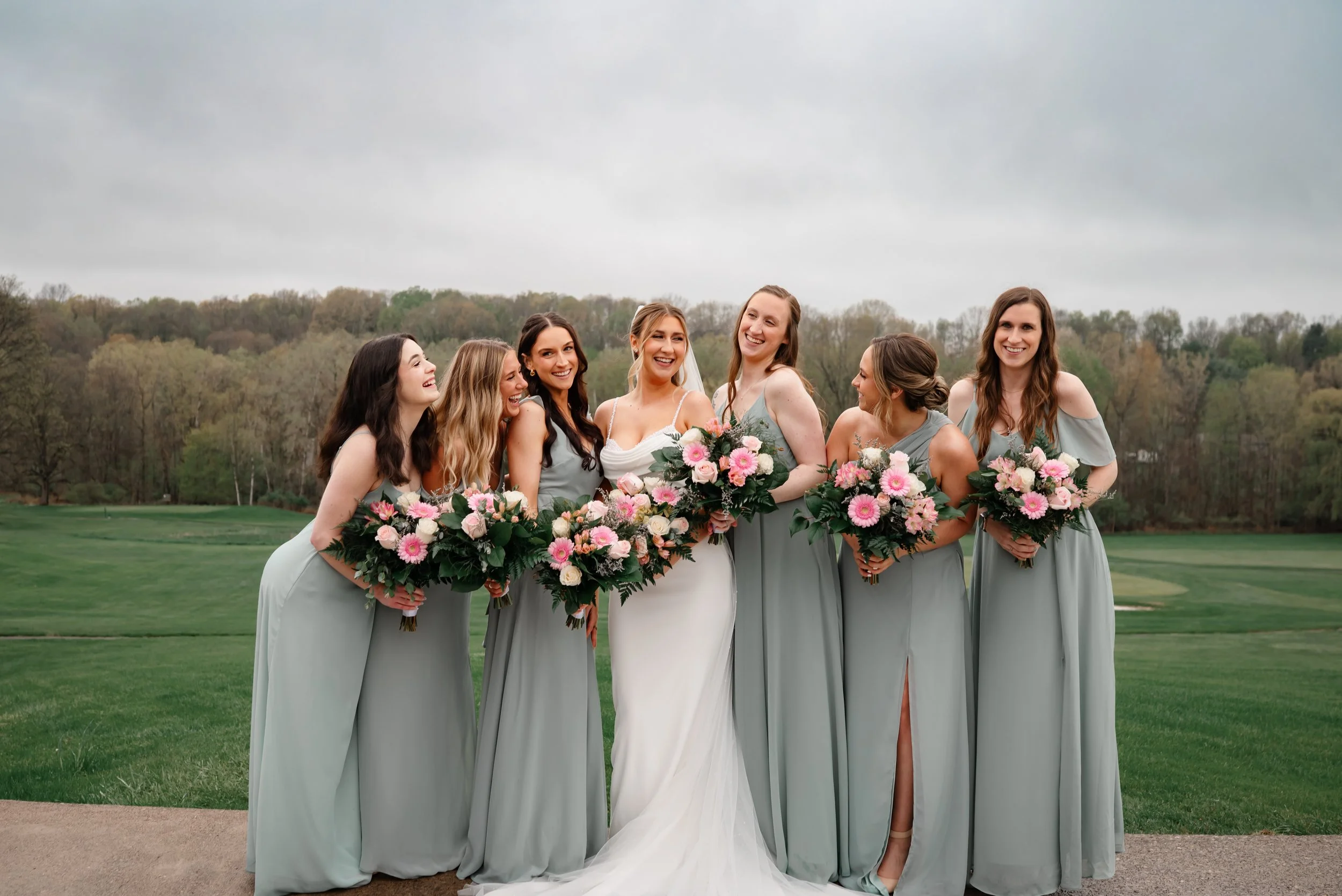 A bride standing with her bridesmaids outdoors during a wedding portrait, all wearing soft green dresses and holding pink and white bouquets, smiling together in a scenic setting.