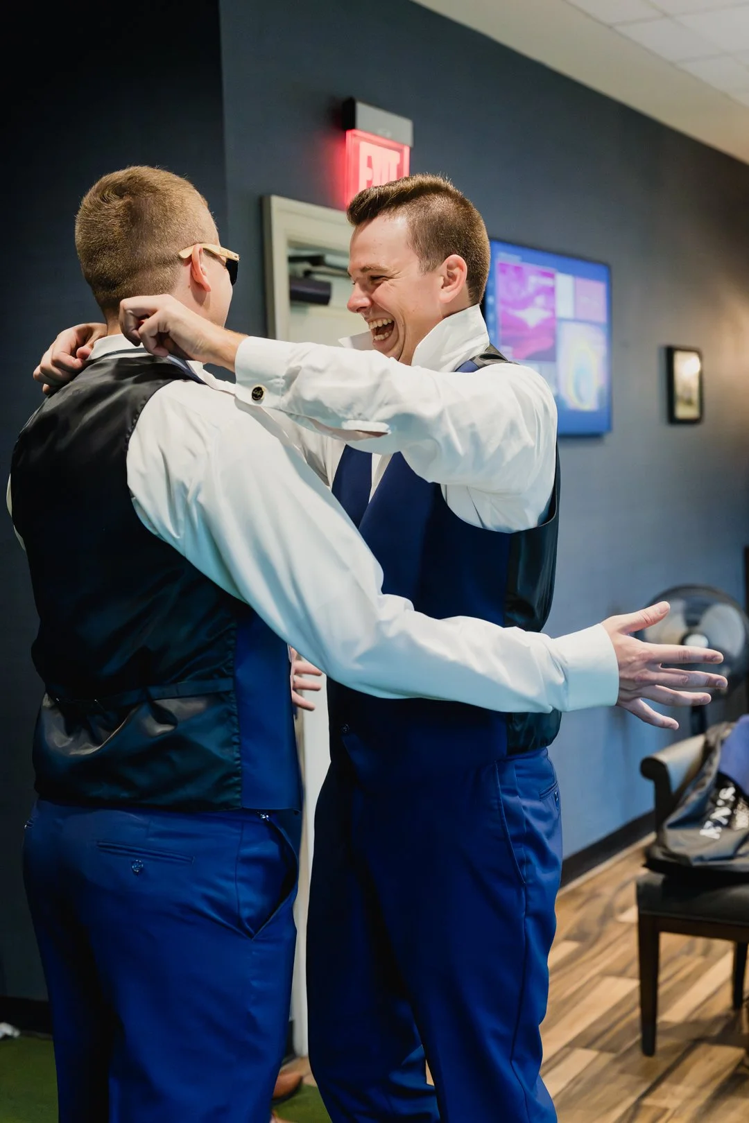 Groom and groomsman laughing and embracing while getting ready for their wedding, wearing matching blue vests and white shirts in an indoor setting.