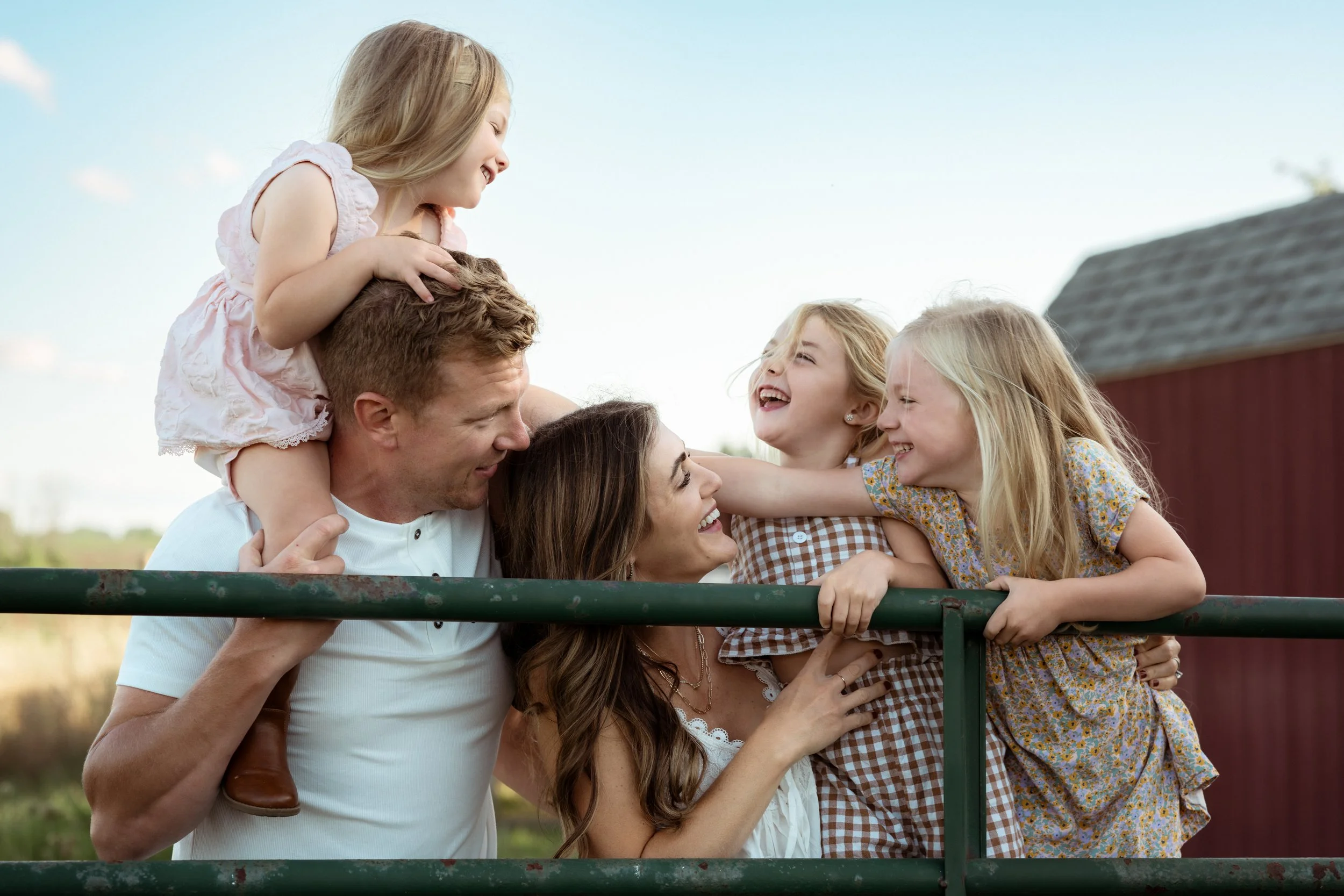 A family of five laughs together over a farm gate during an outdoor family session. Dad holds his youngest daughter on his shoulders while Mom smiles at their three daughters, capturing a joyful, candid moment filled with connection and laughter.