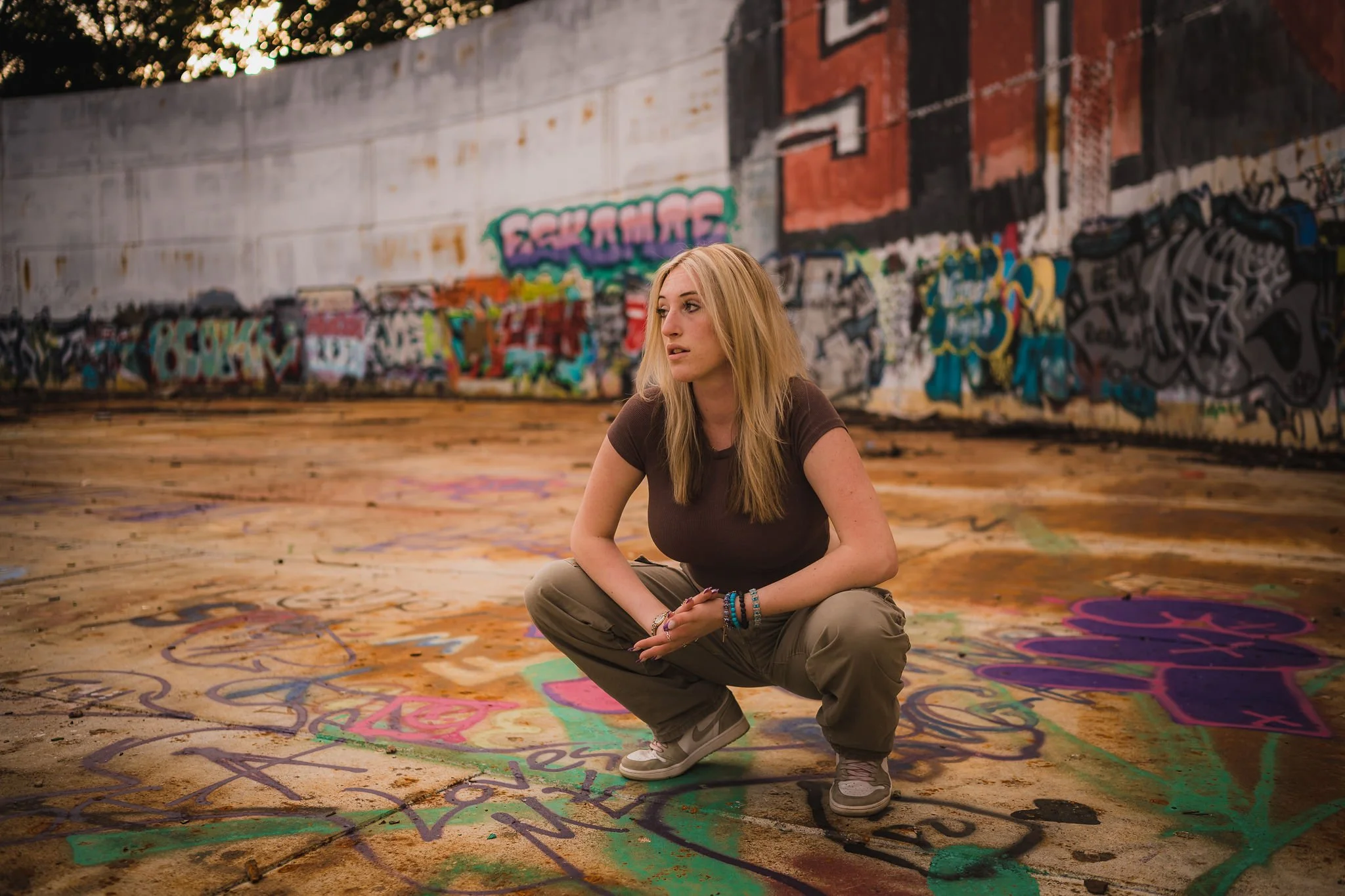 A woman crouching in an urban, graffiti-covered space, wearing casual clothing and looking off to the side, surrounded by colorful street art and textured concrete.
