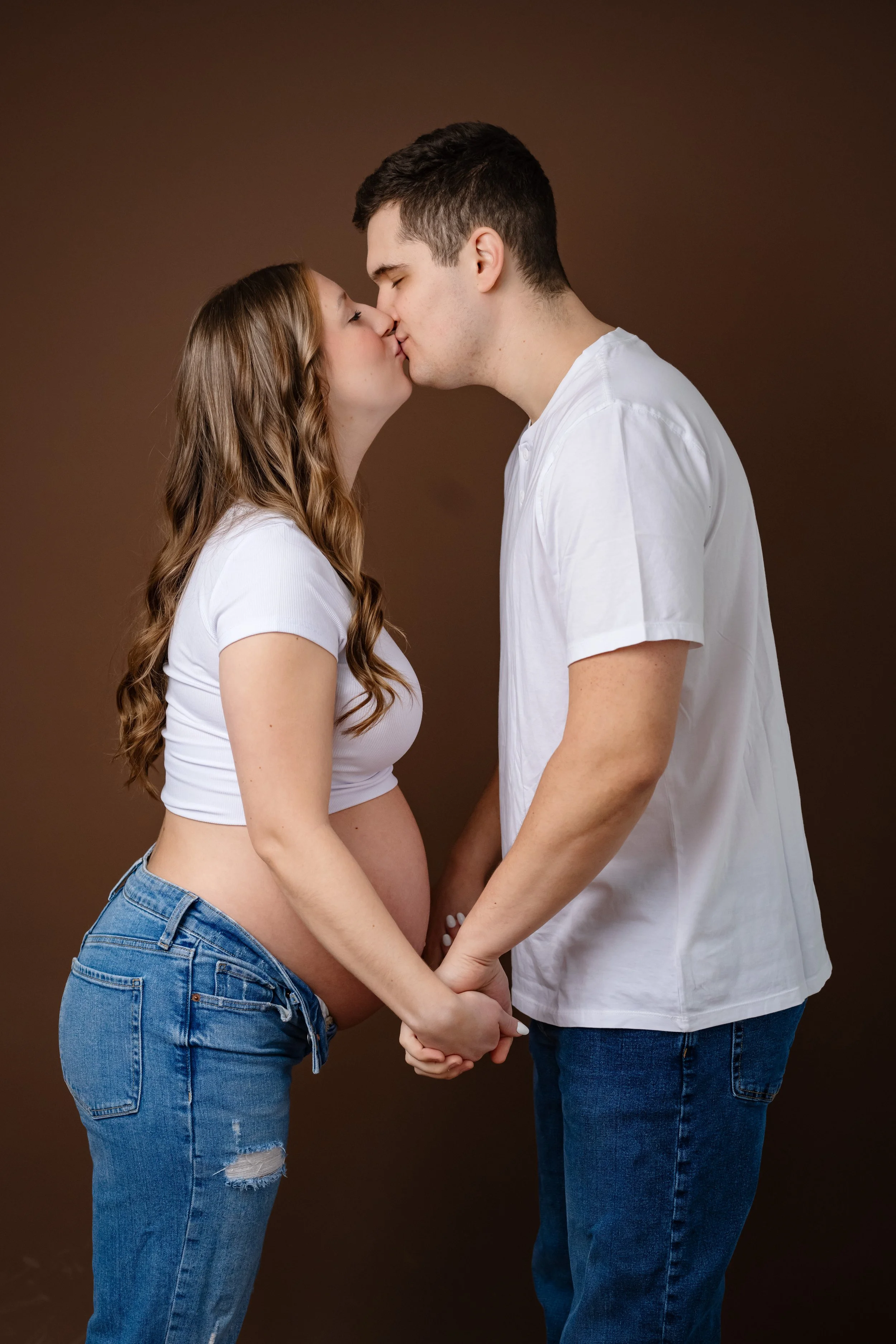 Expectant couple stands face to face, gently holding hands and sharing a kiss, with the mother’s baby bump visible against a warm brown studio backdrop.