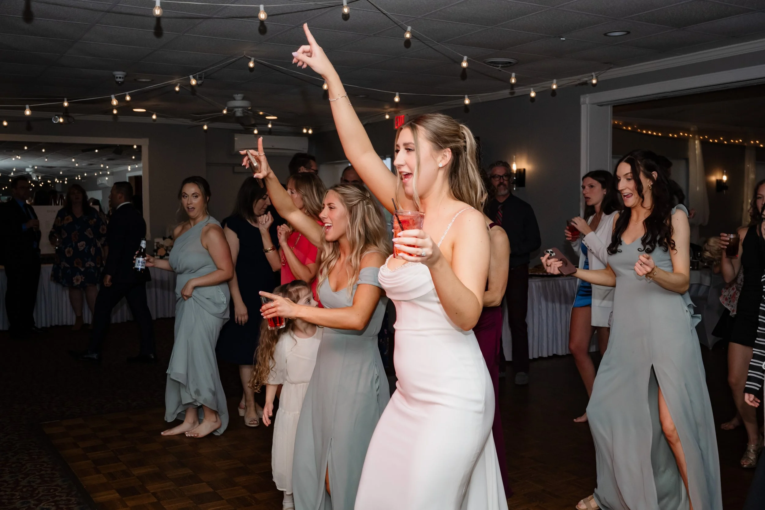 Wedding guests dancing and celebrating on the dance floor during a reception, with bridesmaids raising their arms and holding drinks under warm string lights.