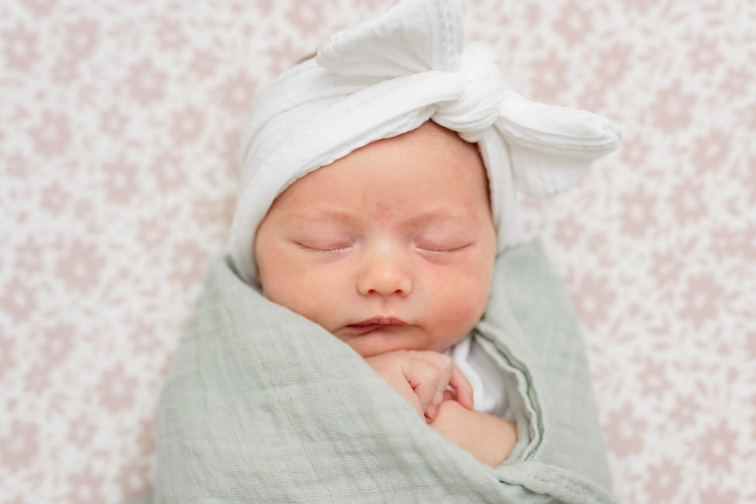 A sleeping newborn baby wrapped in a gray blanket with a white headband tied in a bow on her head, resting peacefully.