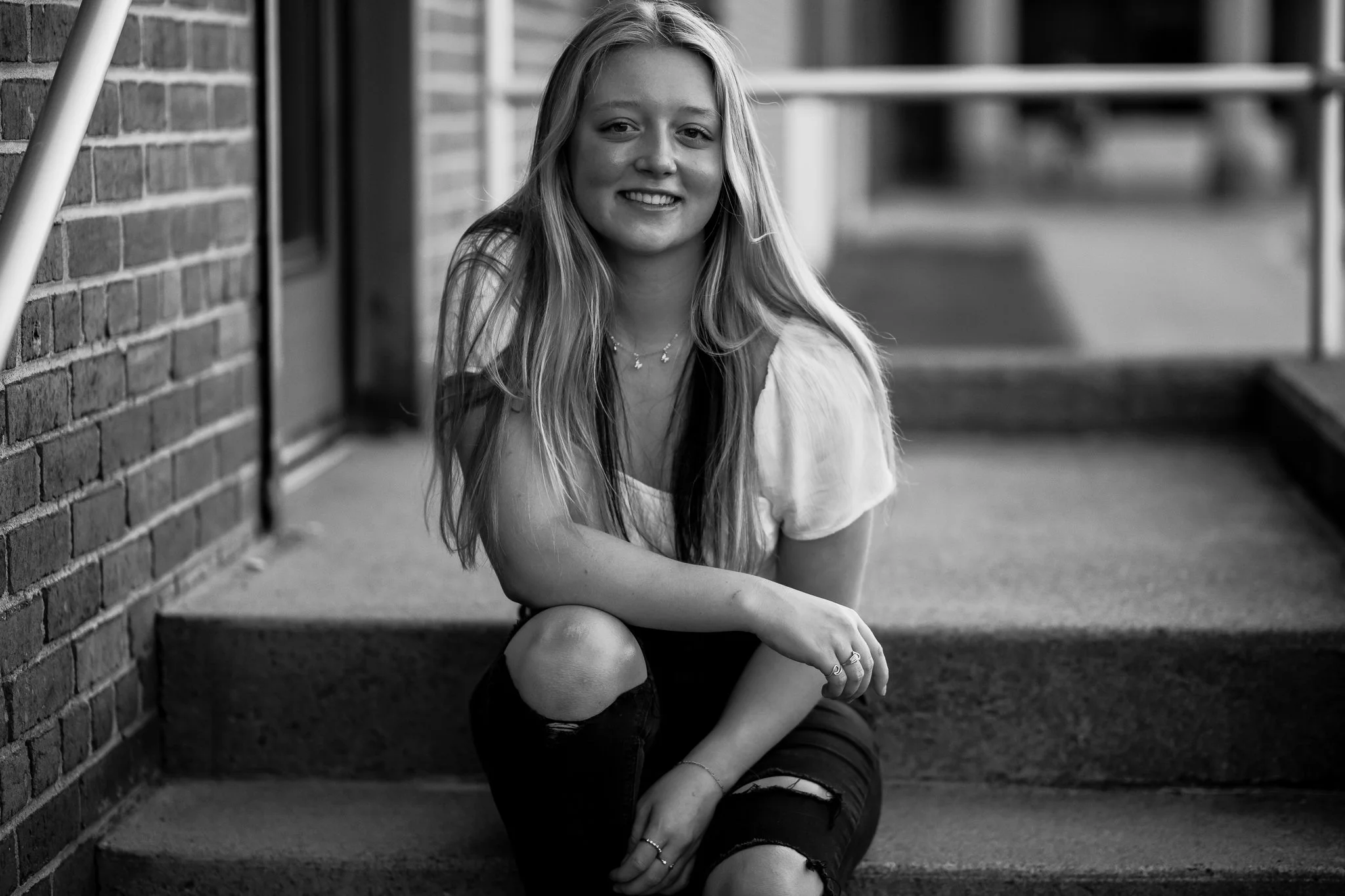 A black-and-white senior portrait of a young woman sitting on concrete steps outdoors, wearing ripped jeans and sneakers, looking at the camera and smiling.