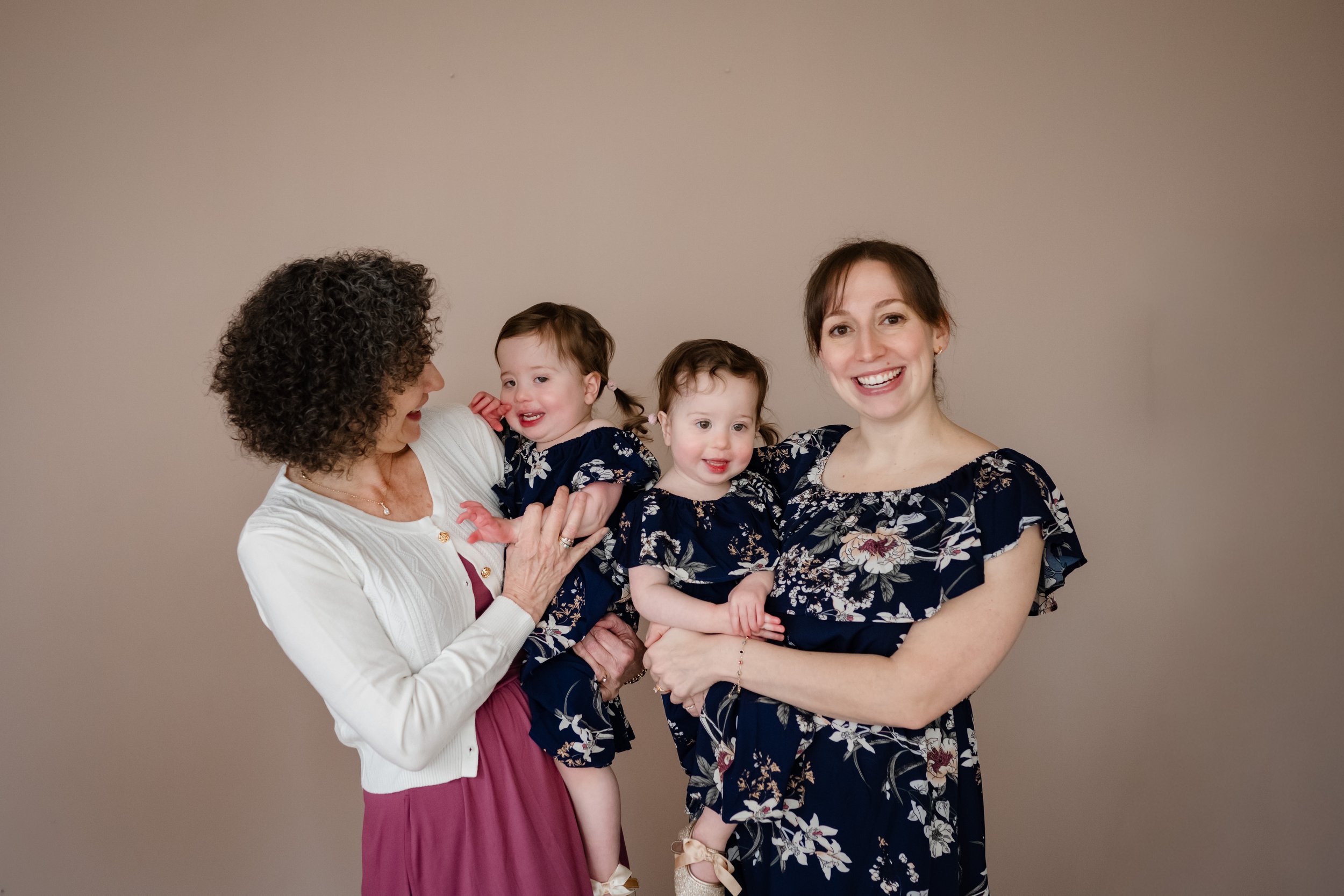 Two women stand side by side holding young twin girls in matching navy floral dresses, smiling and interacting together against a soft neutral backdrop, capturing a joyful, multi-generational family moment.