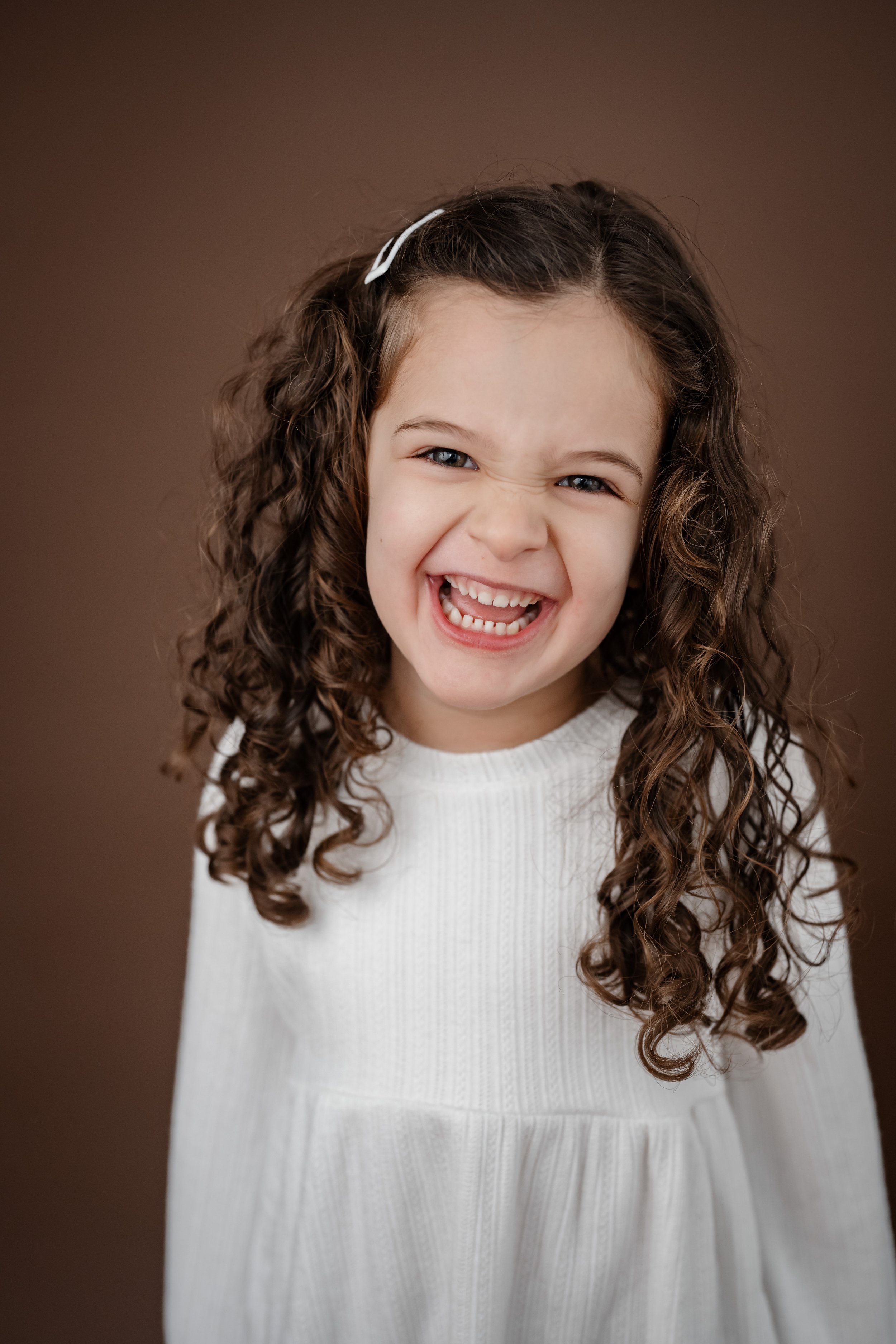 A young child laughing joyfully during a personality portrait session, wearing a white dress with curly hair framing her face against a warm brown backdrop.