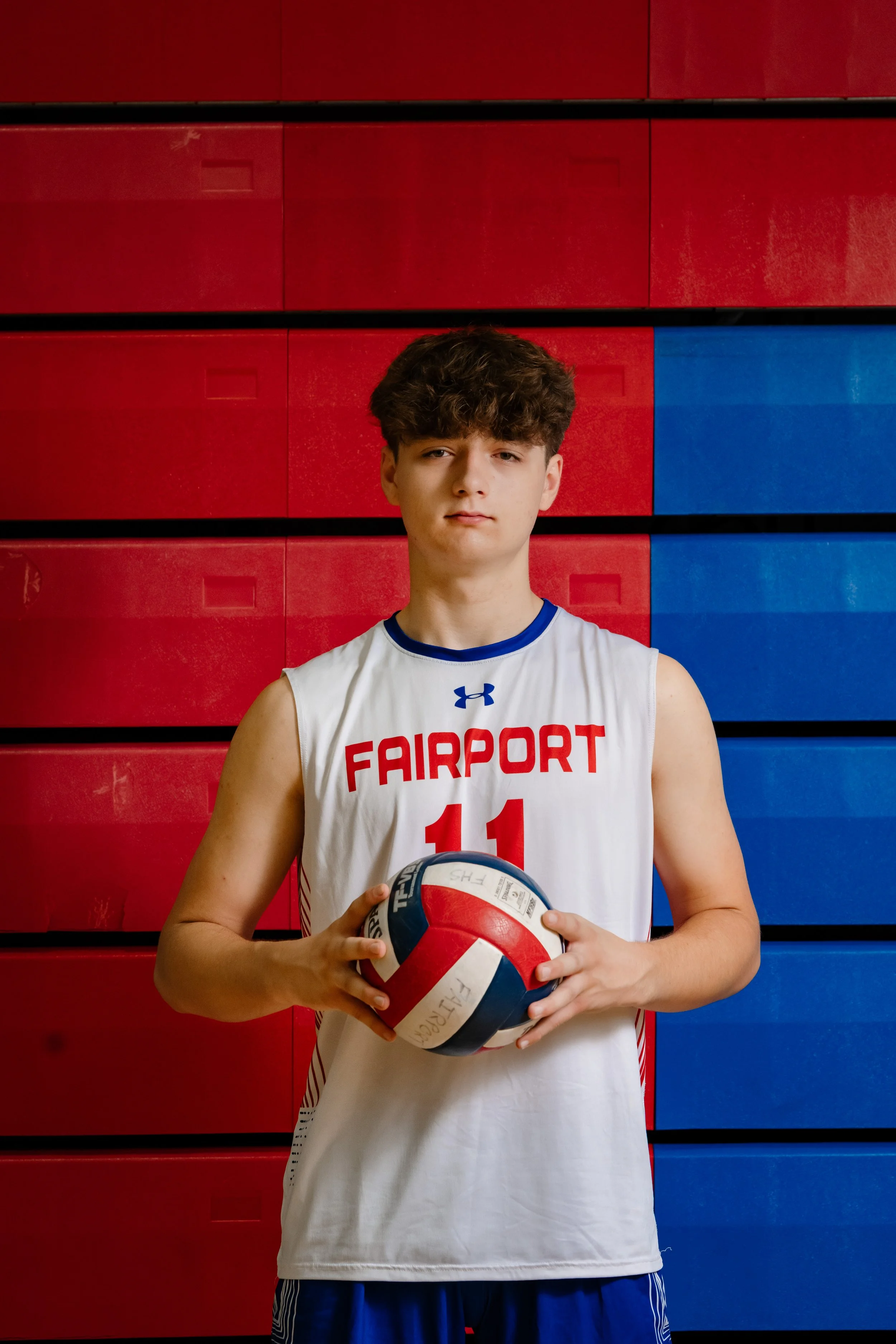 A high school senior athlete standing in a gym holding a volleyball, wearing a Fairport jersey with the number 11, posed in front of red and blue bleachers.