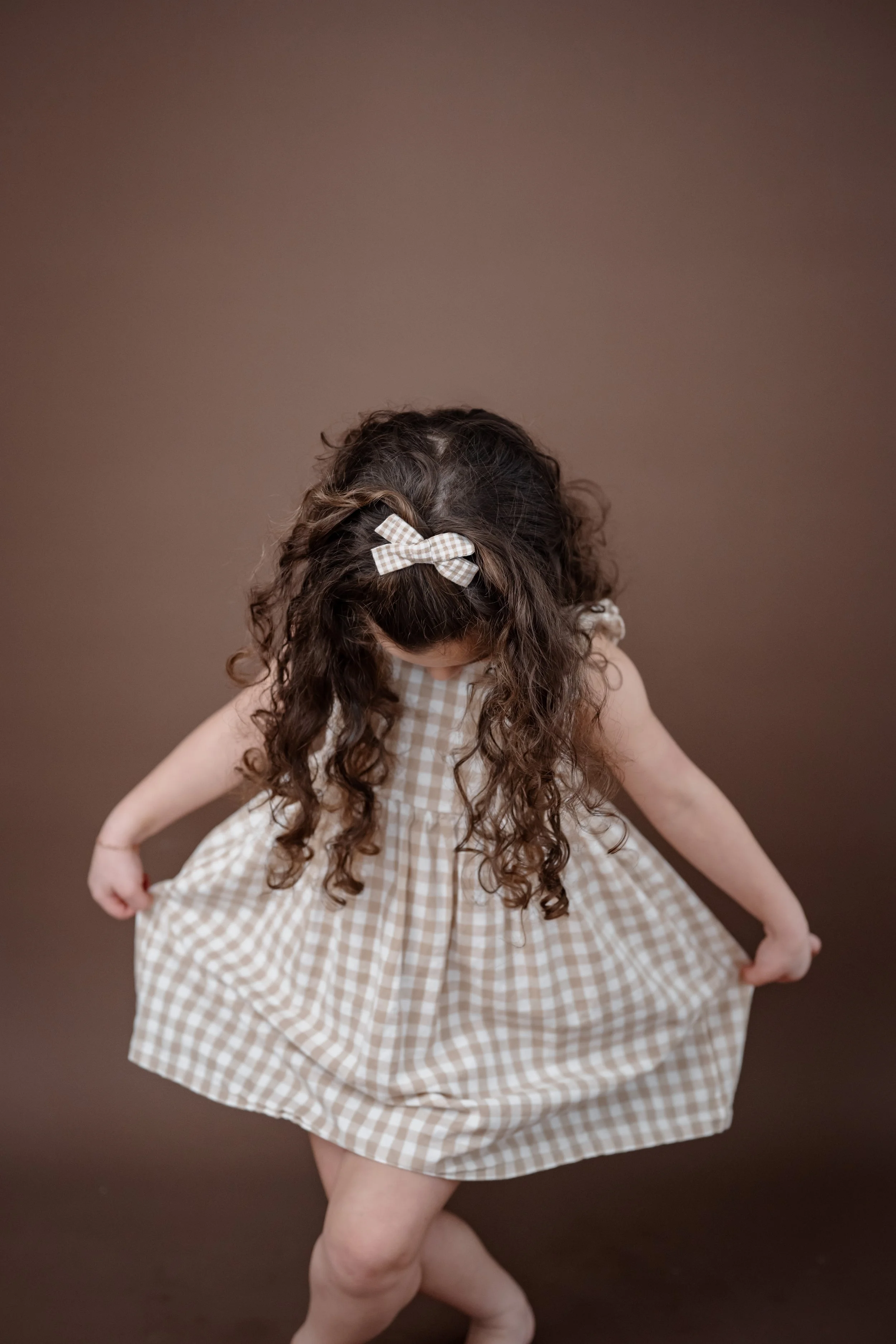 A young girl with curly hair wearing a beige gingham dress and a matching bow, lifting her dress and looking downward against a brown background.
