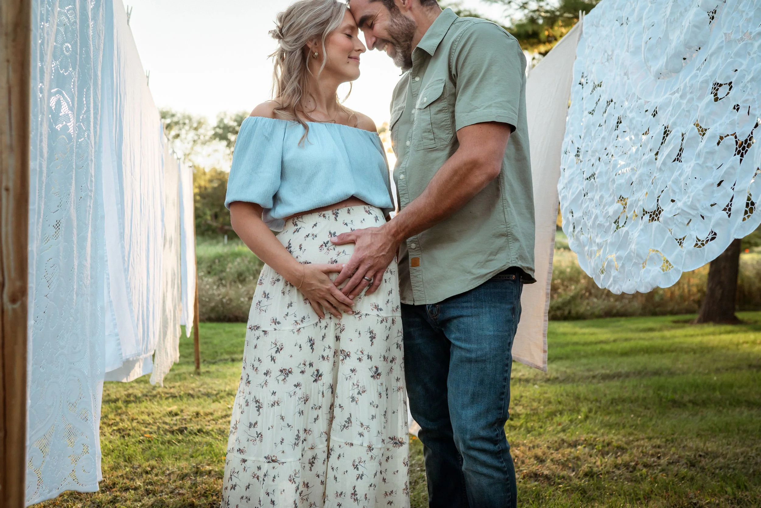 A couple touching her pregnant belly, standing close, with their foreheads touching, outdoors near hanging white and blue fabric decorations.