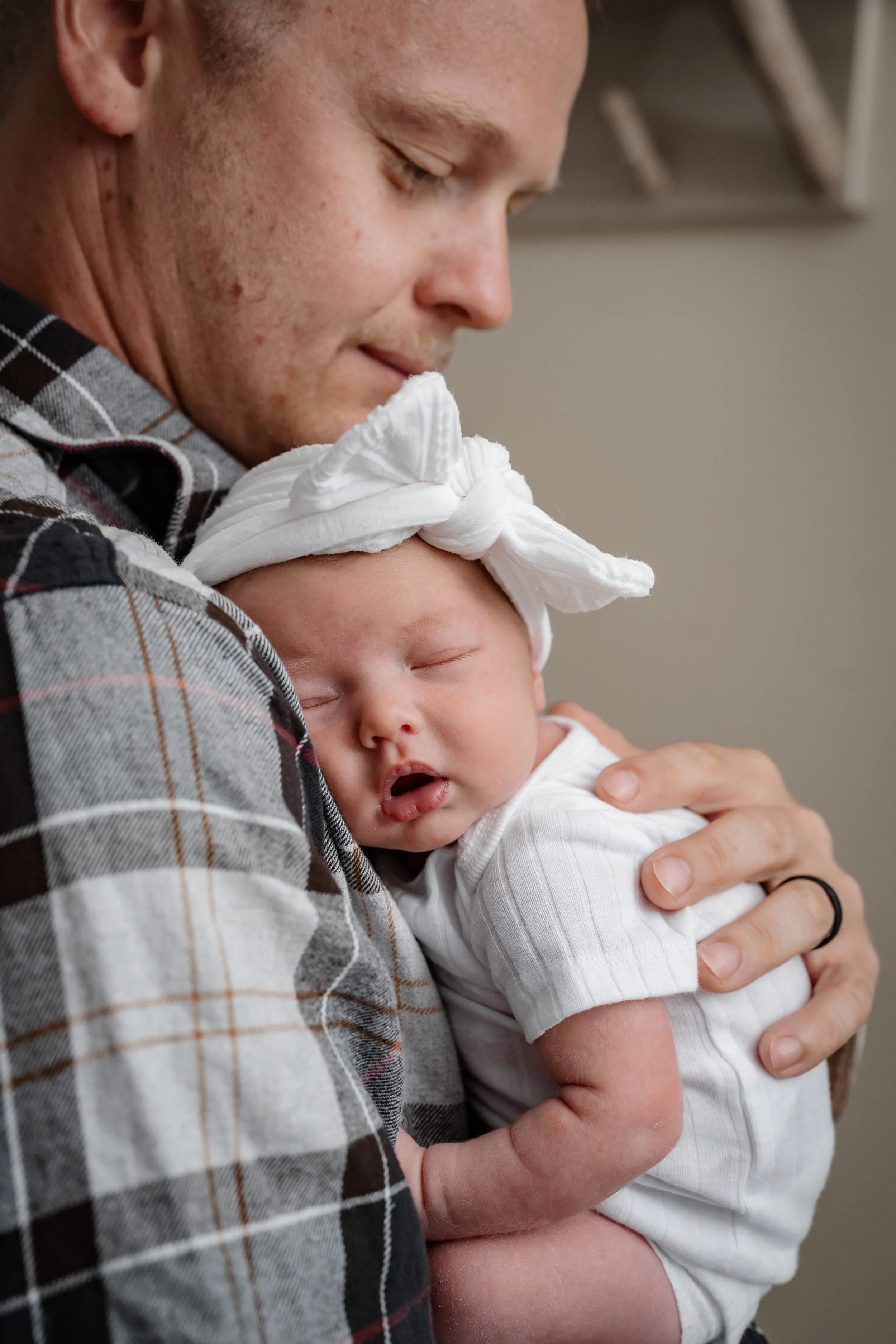 Tender newborn photography of a sleeping baby in a white bow headband, resting peacefully on her father’s chest.
