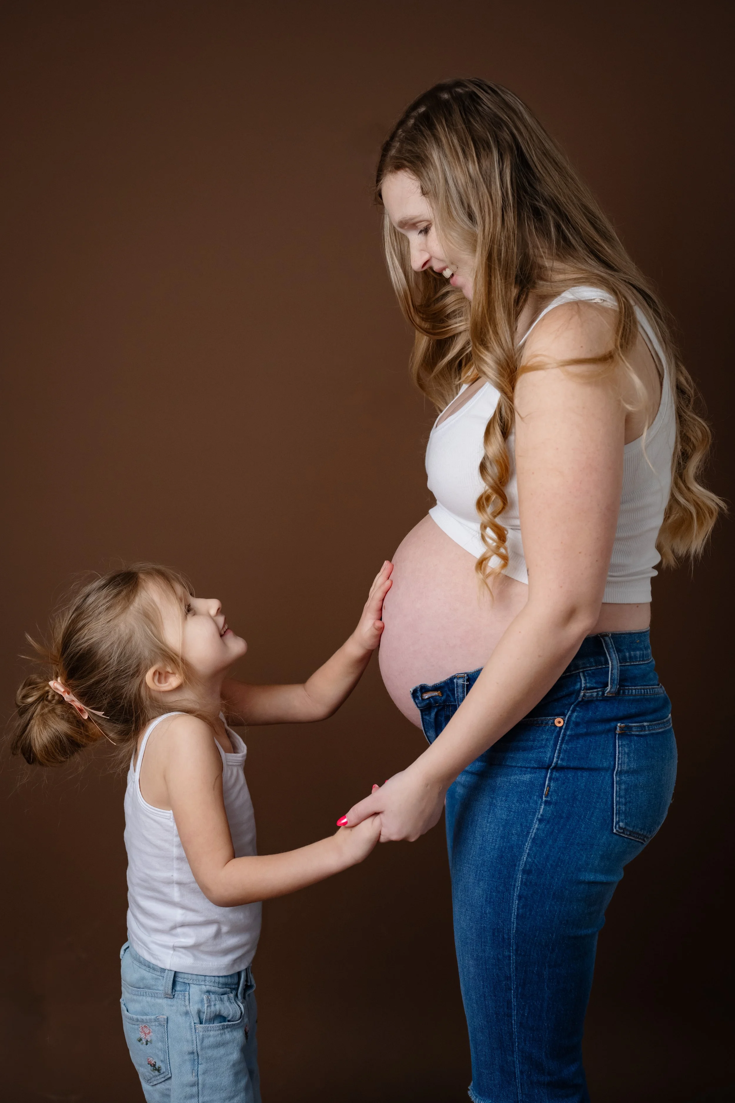 Studio maternity portrait of a pregnant mother and young child, holding hands as the child gently touches her baby bump against a warm brown backdrop.