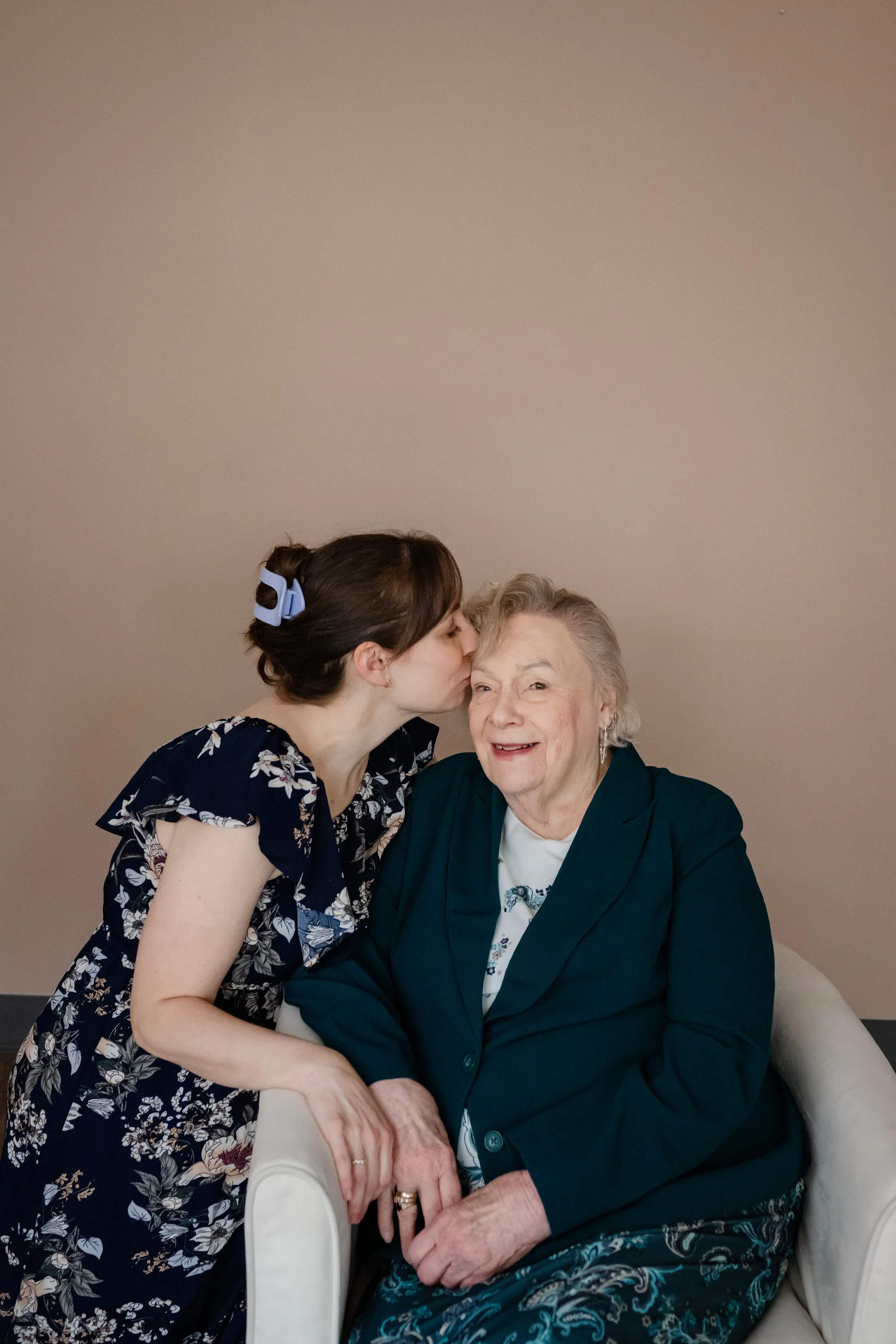 A woman leans in to kiss an elderly woman on the cheek as she sits in a chair, both smiling softly, capturing a tender, multi-generational moment of love and connection.