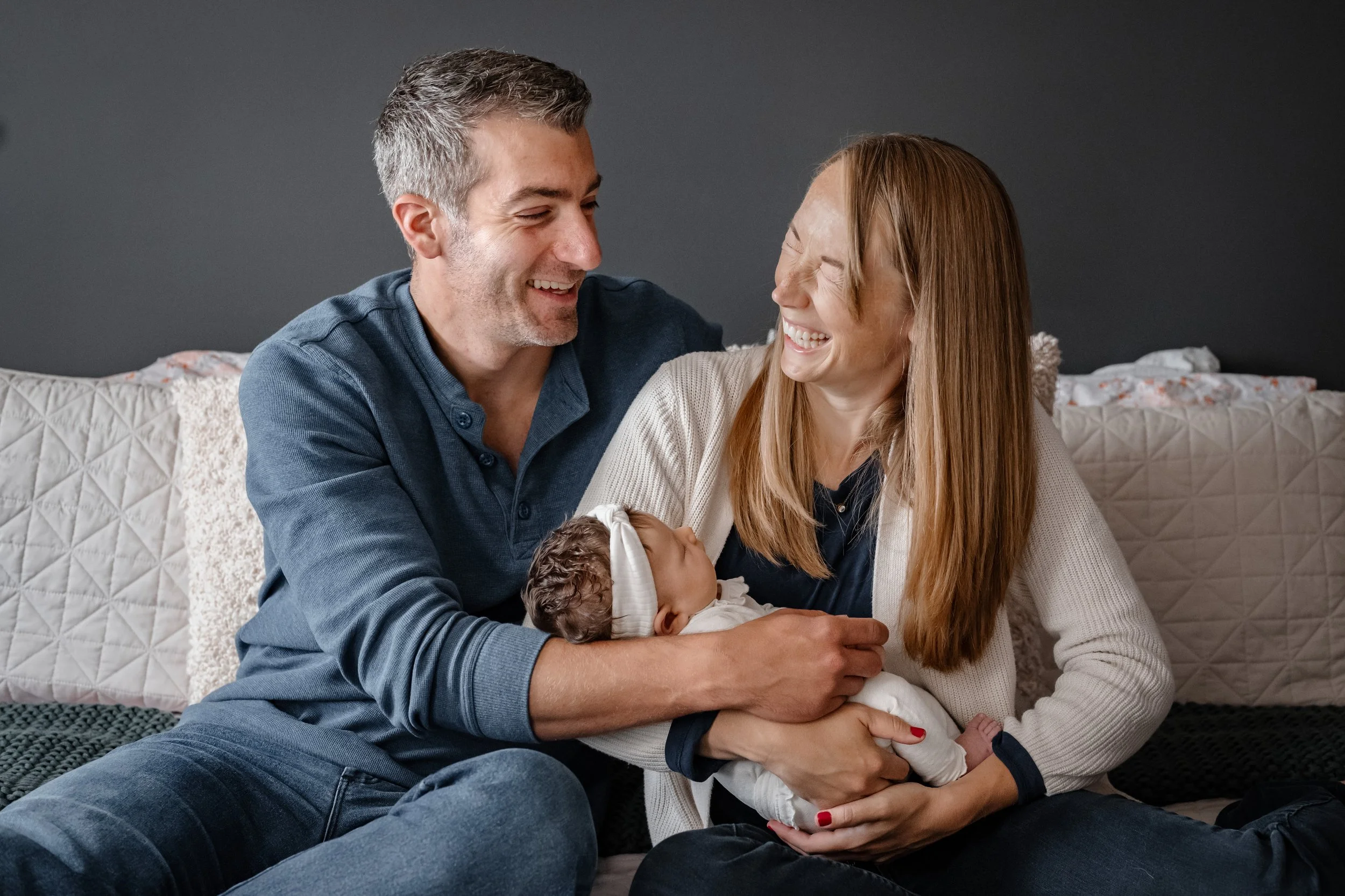 A happy couple with a newborn baby sitting on a bed, smiling at each other.