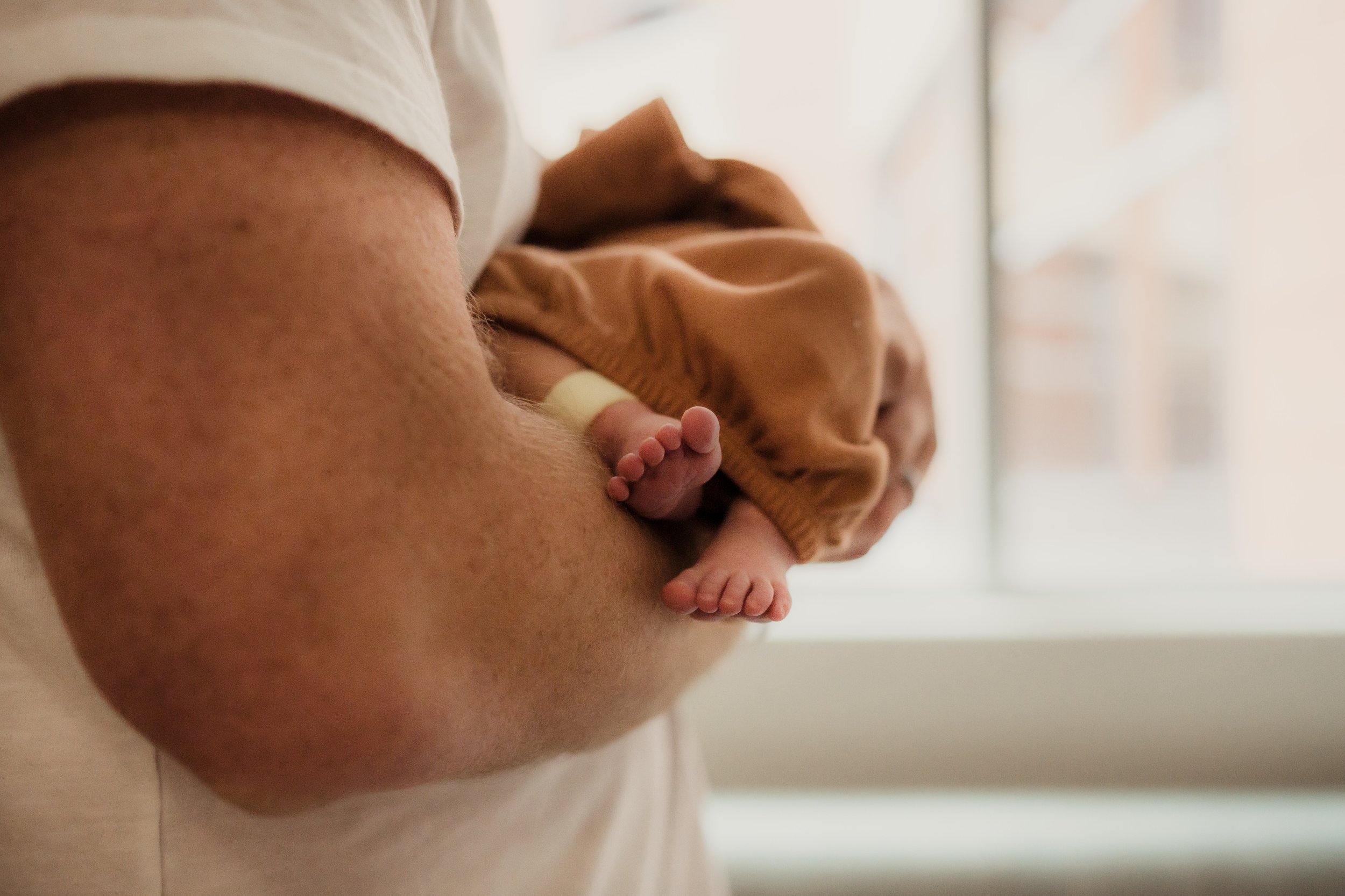 Close-up of a newborn’s tiny feet resting in a parent’s arms near a softly lit window, wrapped in a warm brown blanket.