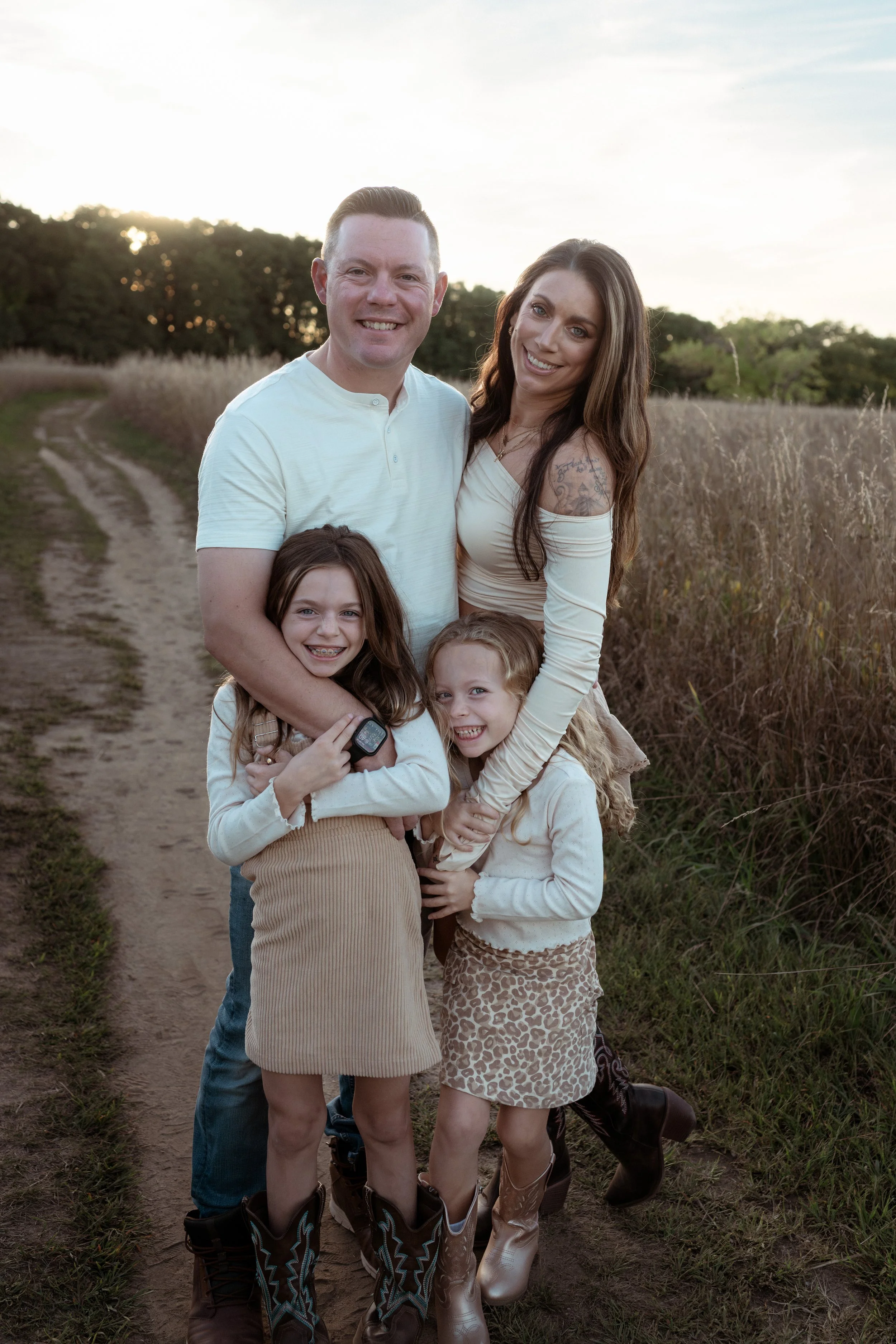 A family of four standing outdoors on a dirt path with tall grasses and trees in the background during sunset. The father and mother are smiling, with the mother wearing a long-sleeved cream top and the father in a white shirt. Two young girls, one holding a watch and the other with a leopard print skirt and cowboy boots, are embracing their parents and smiling.