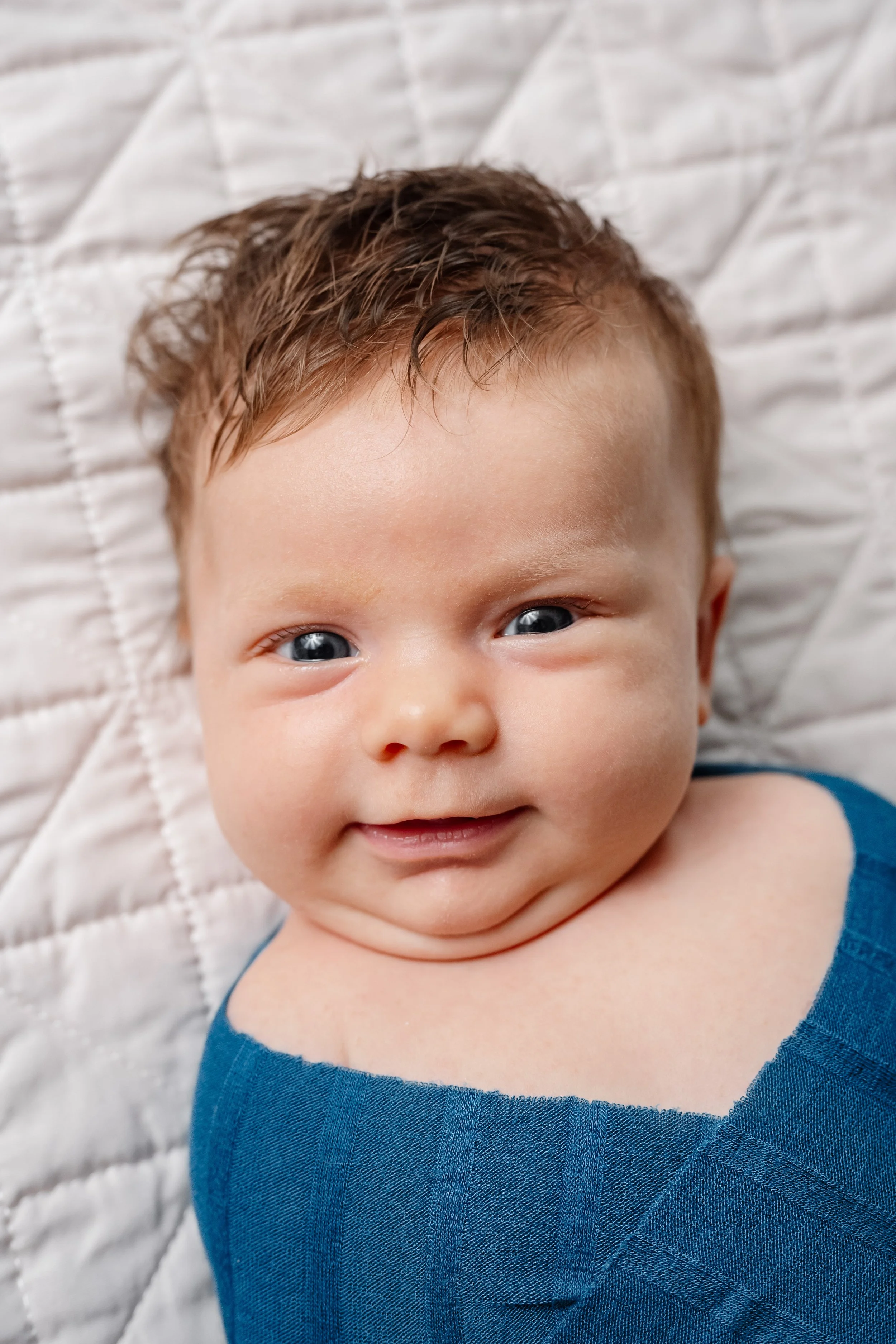 A close-up portrait of a newborn baby lying on a quilted bedspread, wearing a soft blue outfit and looking toward the camera with bright eyes and a gentle smile.