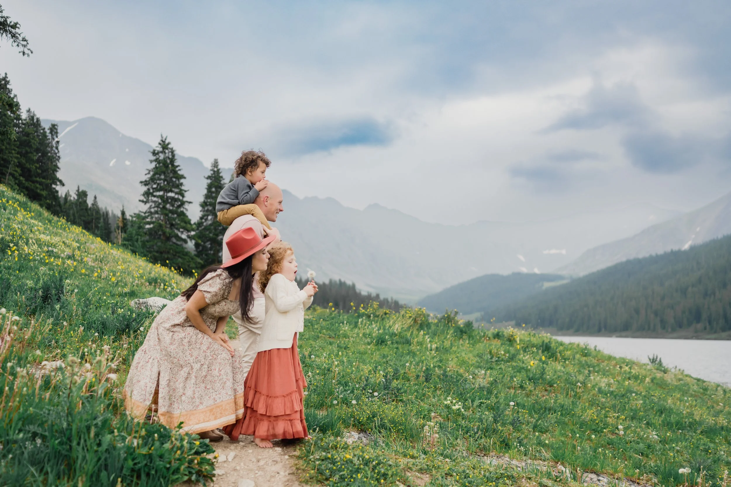 A family standing along a mountain trail during an outdoor photoshoot, with parents crouched beside their children as they look out over wildflowers, evergreen trees, and a lake in the distance. Soft mountain light and dramatic clouds create a warm, 