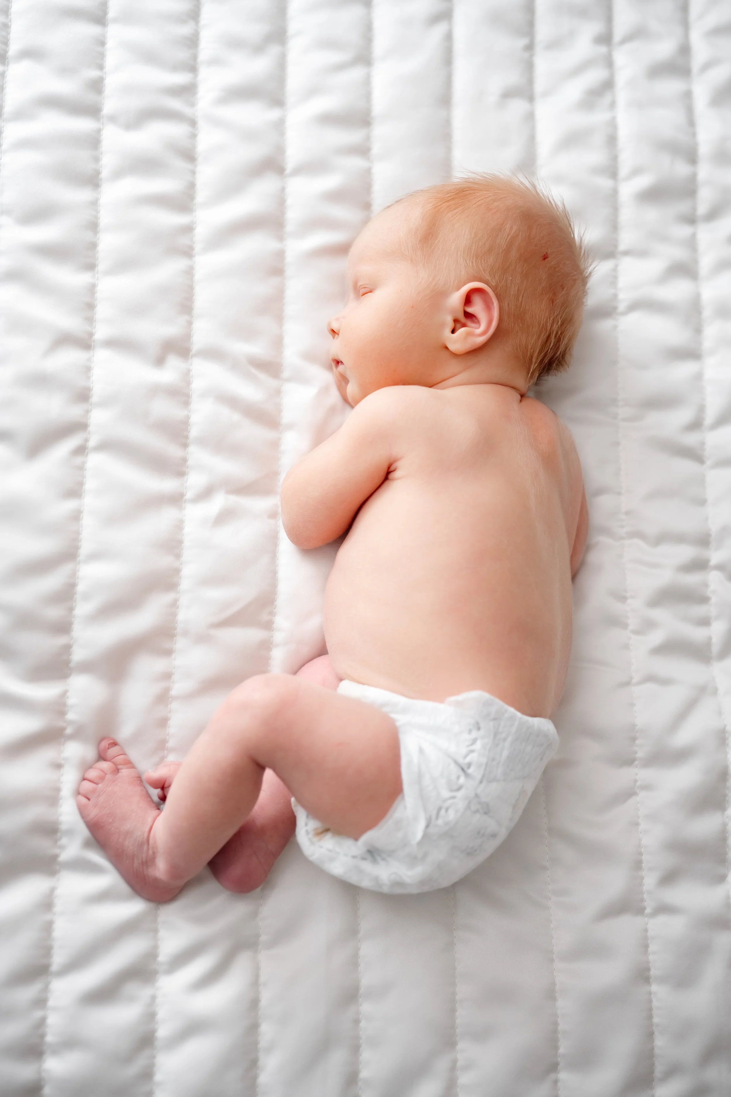 Overhead view of a newborn baby sleeping on a white quilted blanket, wearing a diaper, captured during a soft, natural in-home newborn photography session.