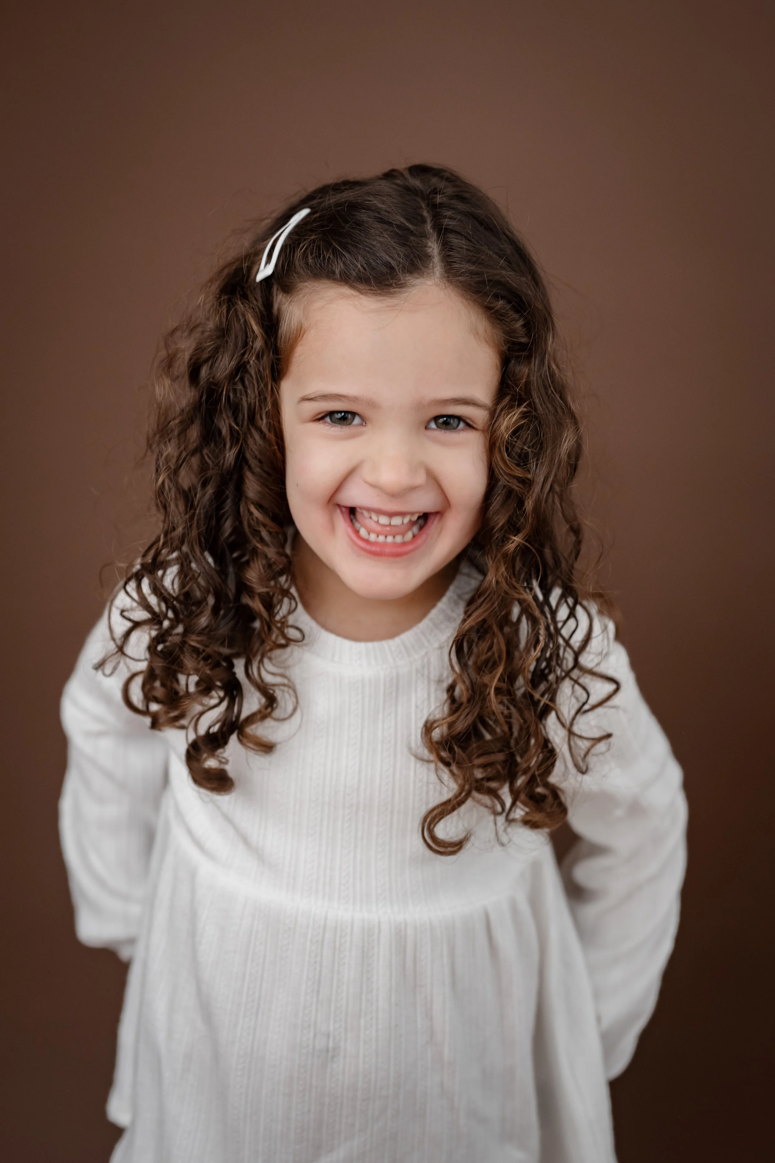 Portrait of a young girl with curly brown hair, wearing a white dress, smiling against a brown background.